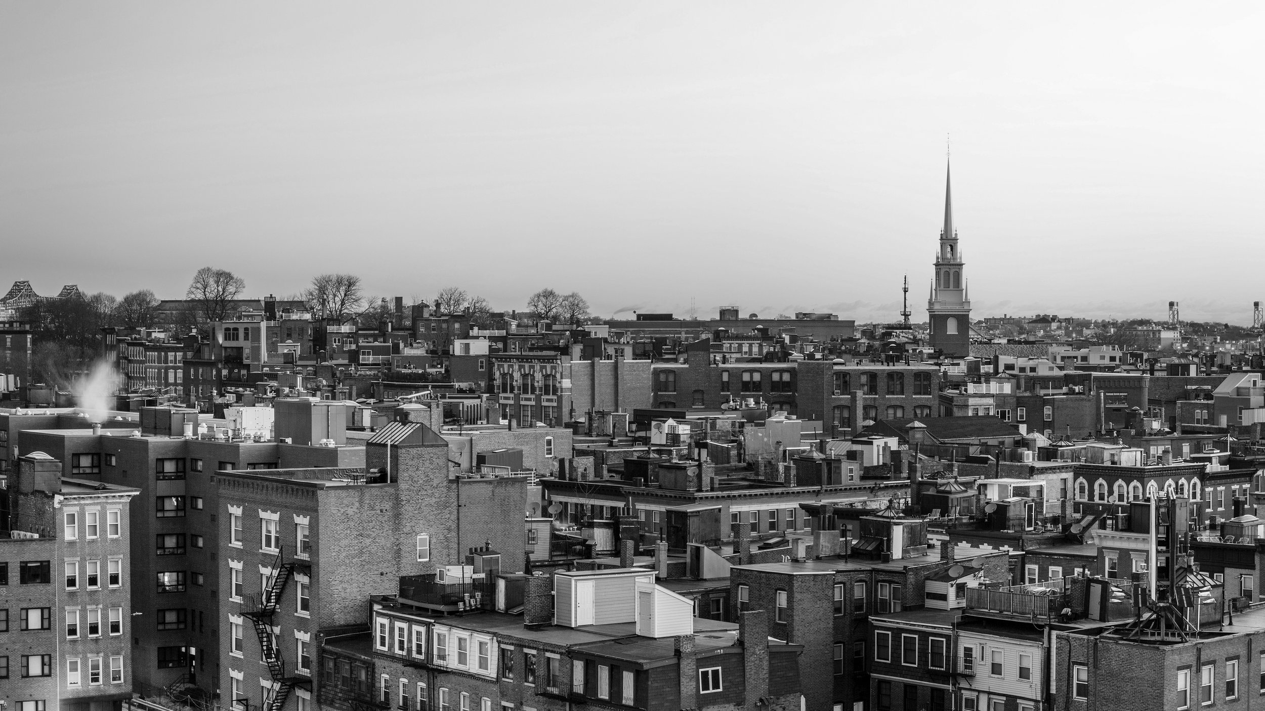 Black and white photo of a city skyline with densely packed buildings and a prominent church steeple, with a bridge and water in the distance.