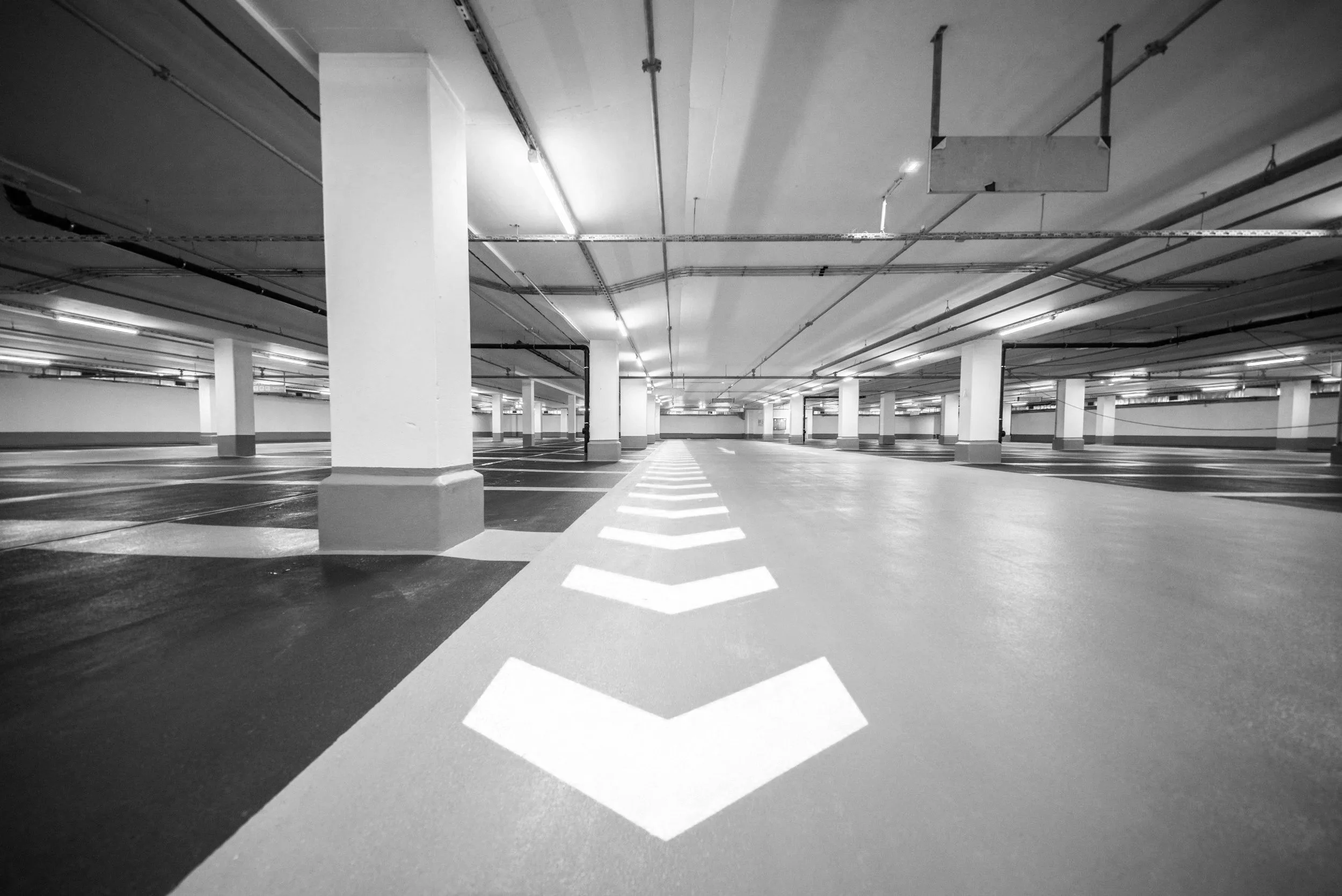 Empty indoor parking garage with white painted arrows on the floor indicating multiple parking spaces, supported by white columns and a ceiling with pipes and lights.