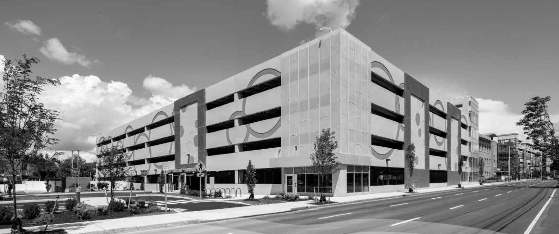 A modern multi-story parking garage with circular artwork on its exterior, empty street, trees, and a partly cloudy sky.