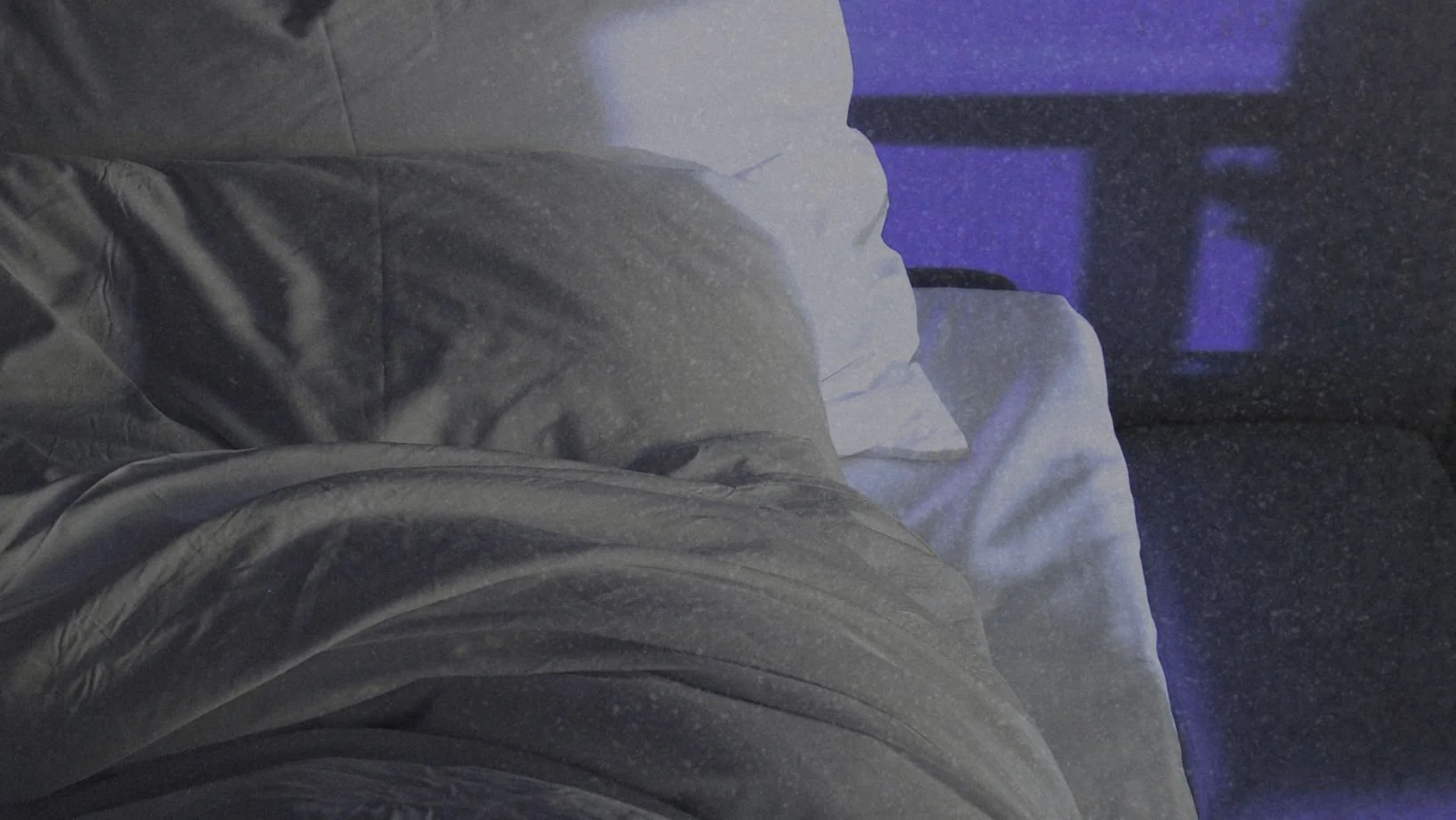 Unmade bed in a dark bedroom illuminated by soft purple light from a window.
