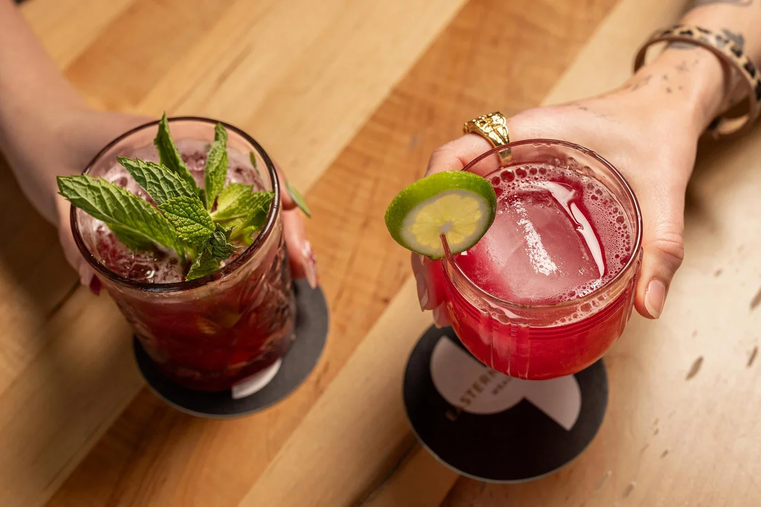 Two cocktails, one garnished with fresh mint leaves and the other with a lime wheel, on coasters on a wooden table.