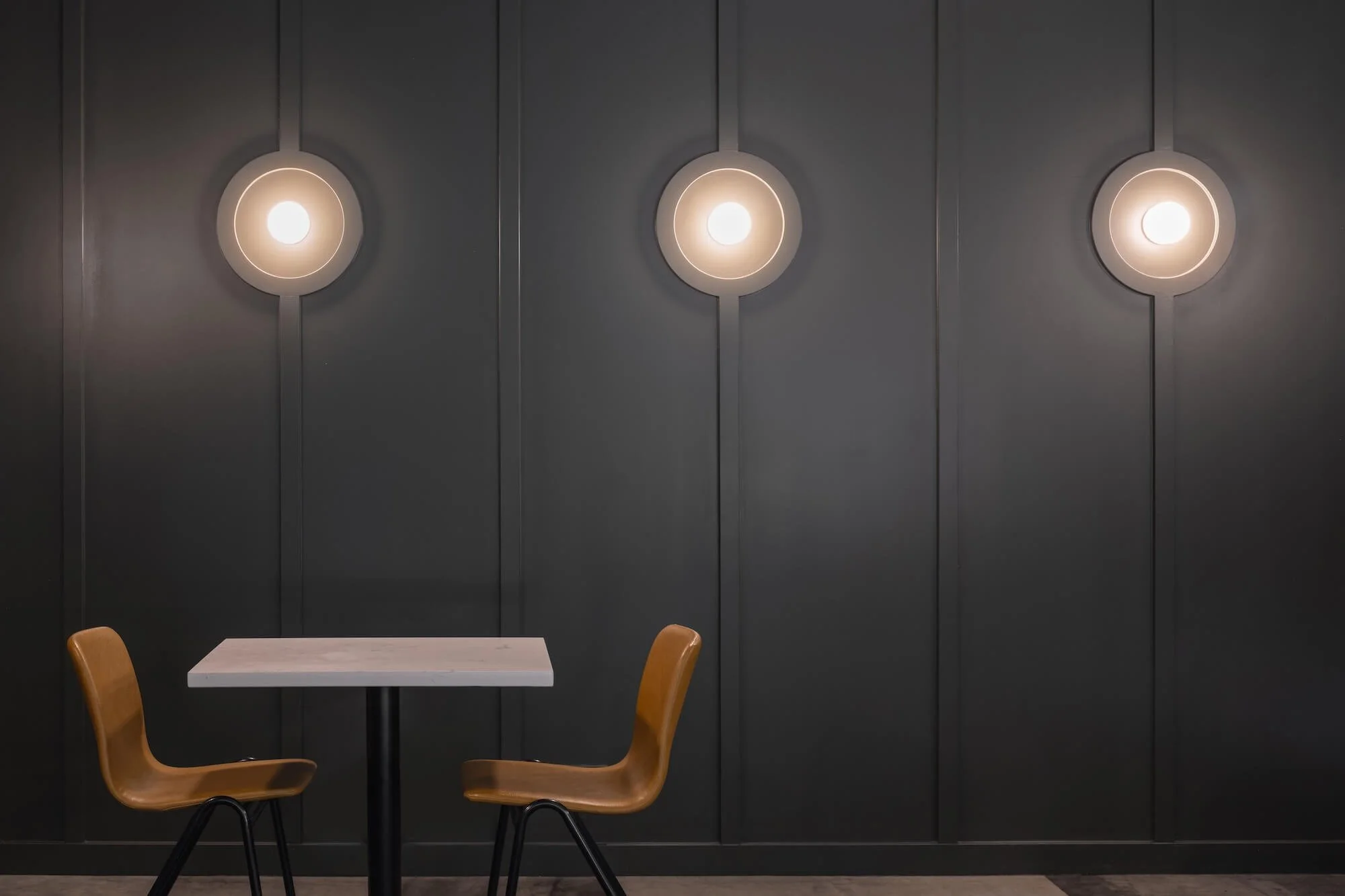 A small table with two wooden chairs in front of a gray wall with three bright circular lights mounted on it.