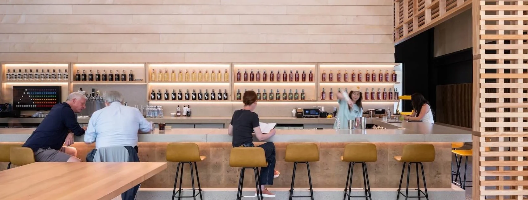 A modern bar with a light wood counter, three yellow bar stools, and four people sitting and working or chatting. The back wall features shelves with bottles of various colors and sizes, a color chart, and bar equipment. A woman behind the counter is shaking a cocktail shaker, another woman appears to be using a blender or coffee machine, and two men are seated at the bar, engaged in conversation.