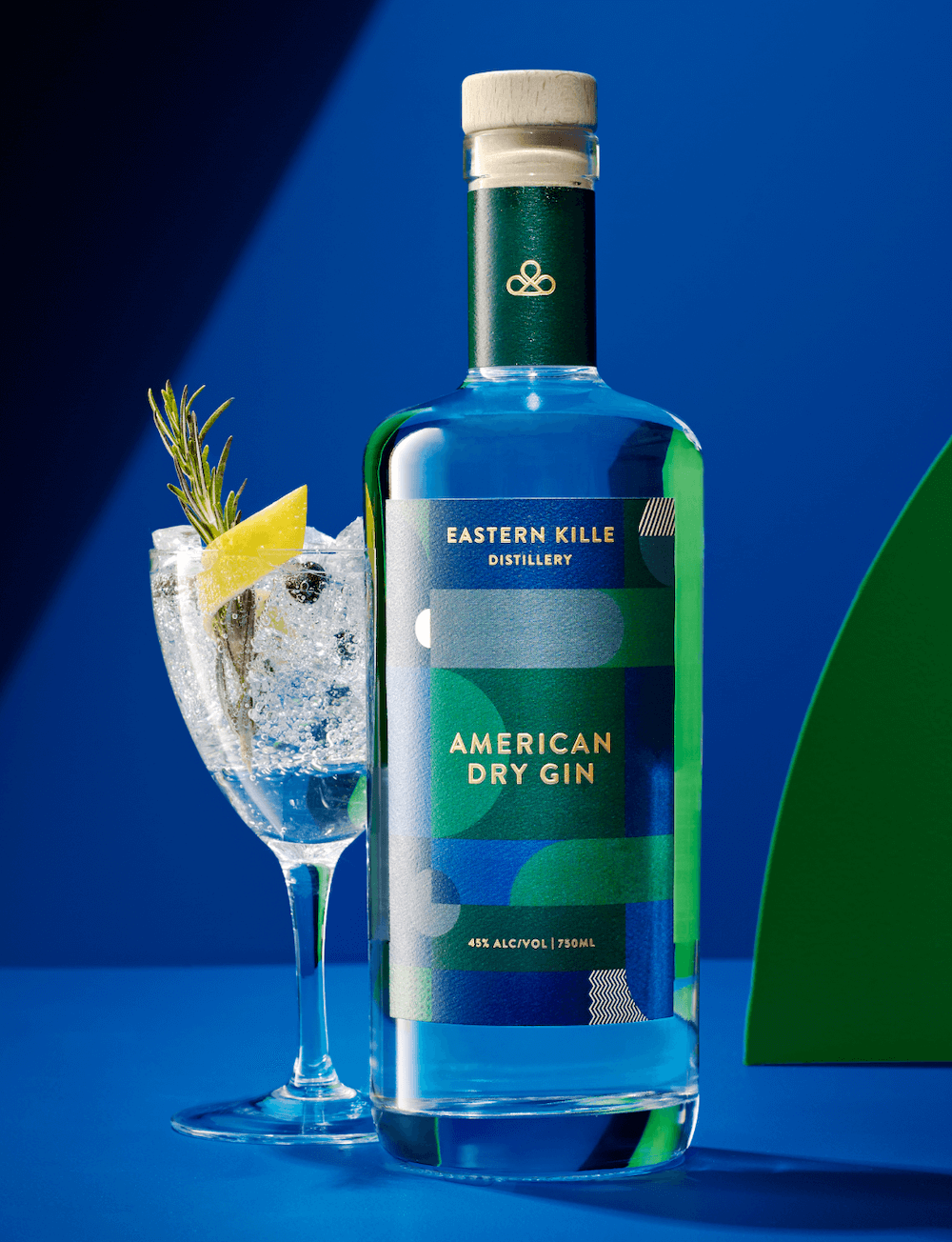 A bottle of Eastern Kille American Dry Gin with a glass of gin and tonic garnished with lemon and rosemary on ice, set against a blue background.