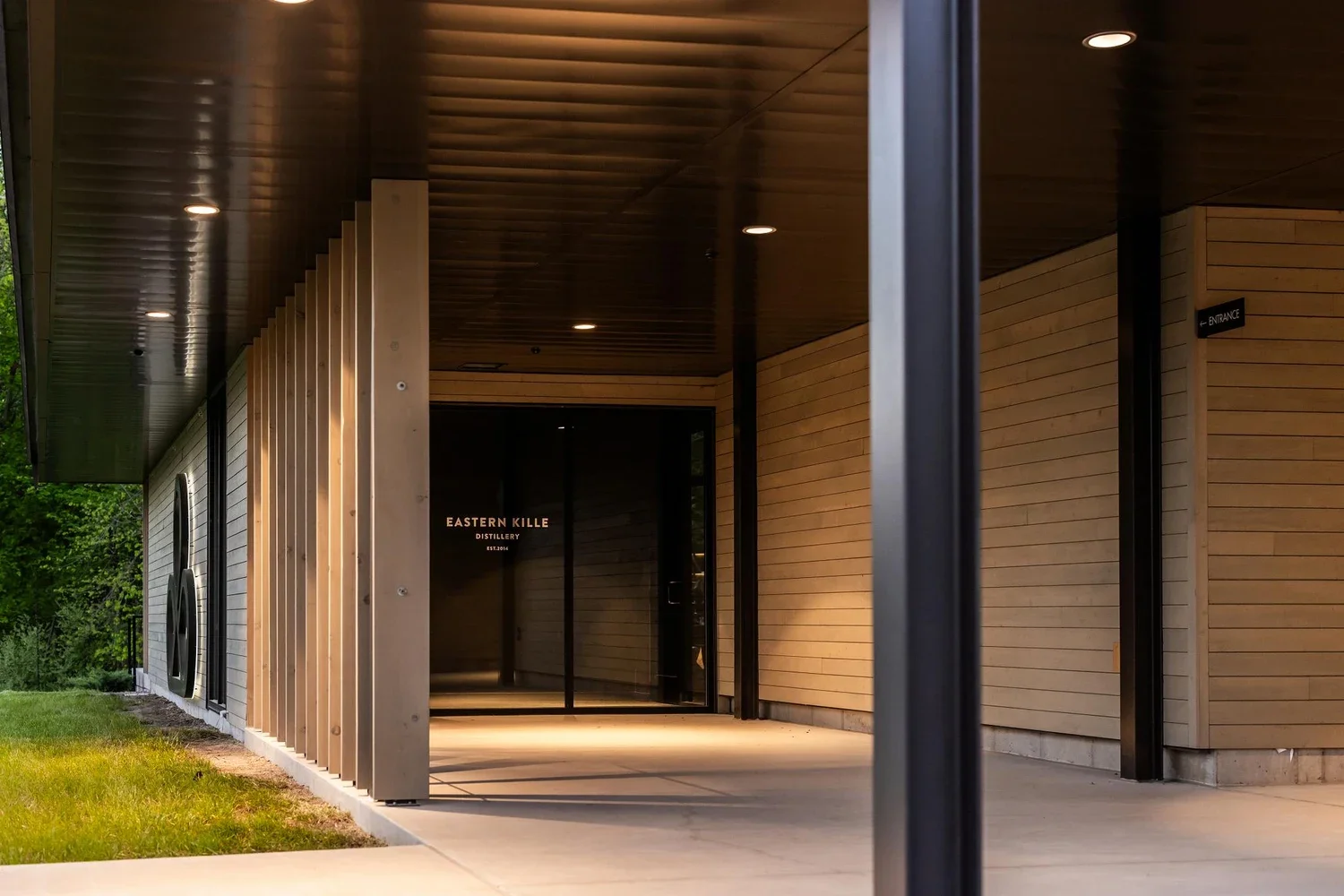 Exterior view of a modern building with a wooden facade, black metal supports, and a partially covered entrance. A sign reads 'Eastern Kille Distillery' near the entrance door.