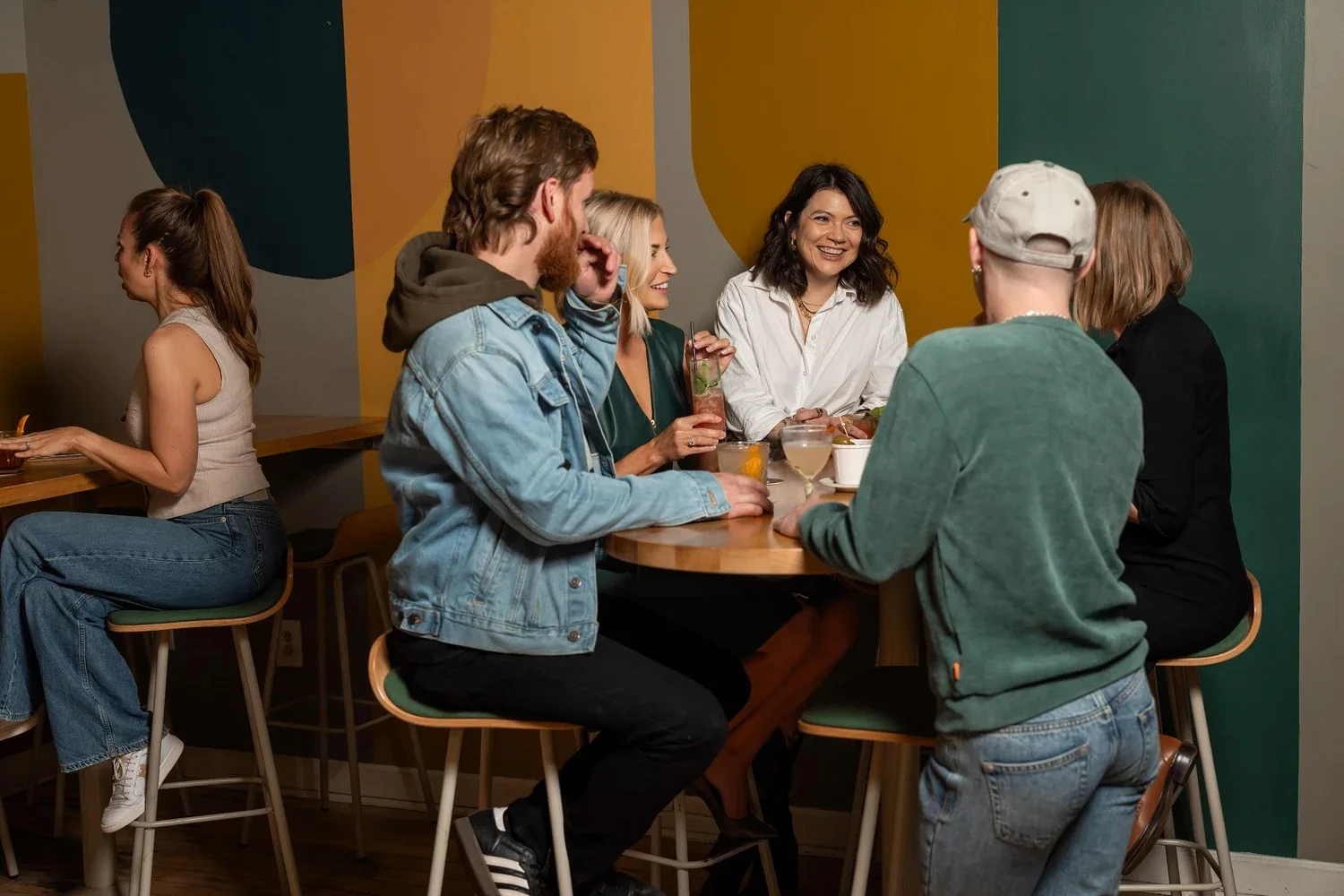 Group of friends socializing at a bar or restaurant, sitting around a circular table with drinks, laughing and talking.