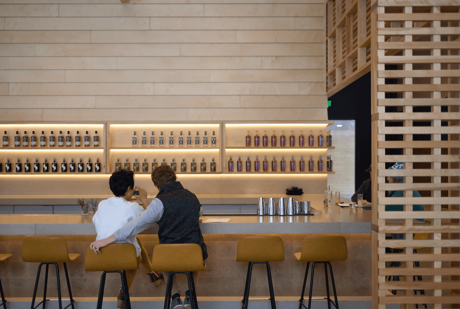 Two people sitting on barstools at a counter in a modern, minimalist bar or cafe, engaging in conversation. Behind them, a wall features illuminated shelves with bottles arranged in a pattern. The setting has light wood paneling and a wooden lattice partition.