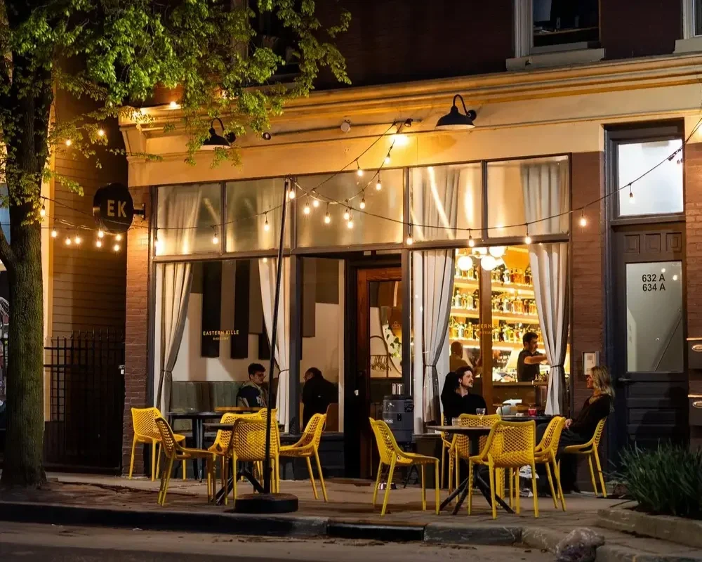 People sitting and dining outside a restaurant at night with string lights and warm lighting inside, yellow chairs, and a visible bar shelf inside.
