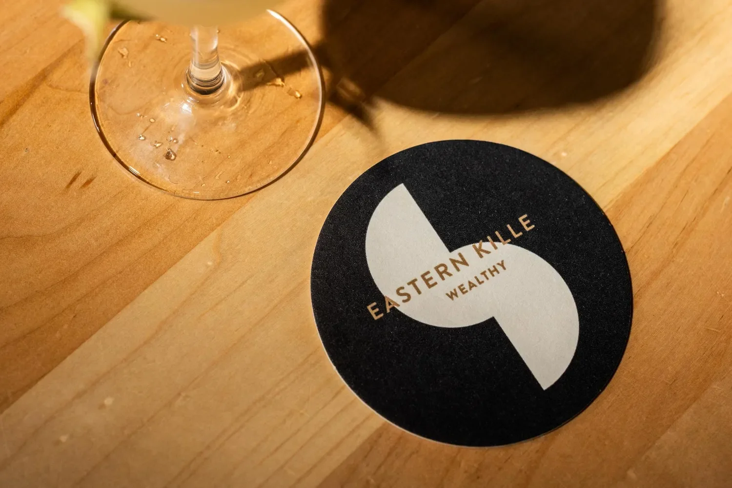 A wine glass with some water droplets on the base, placed on a wooden surface next to a round black and white coaster with the words 'Eastern Kille Wealthy' on it.