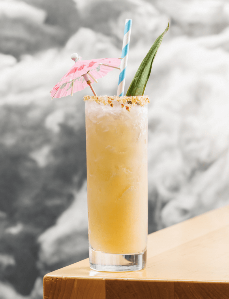 A tall glass of tropical cocktail with a pink paper umbrella, a blue and white striped straw, and a pineapple leaf garnish, placed on a wooden surface.
