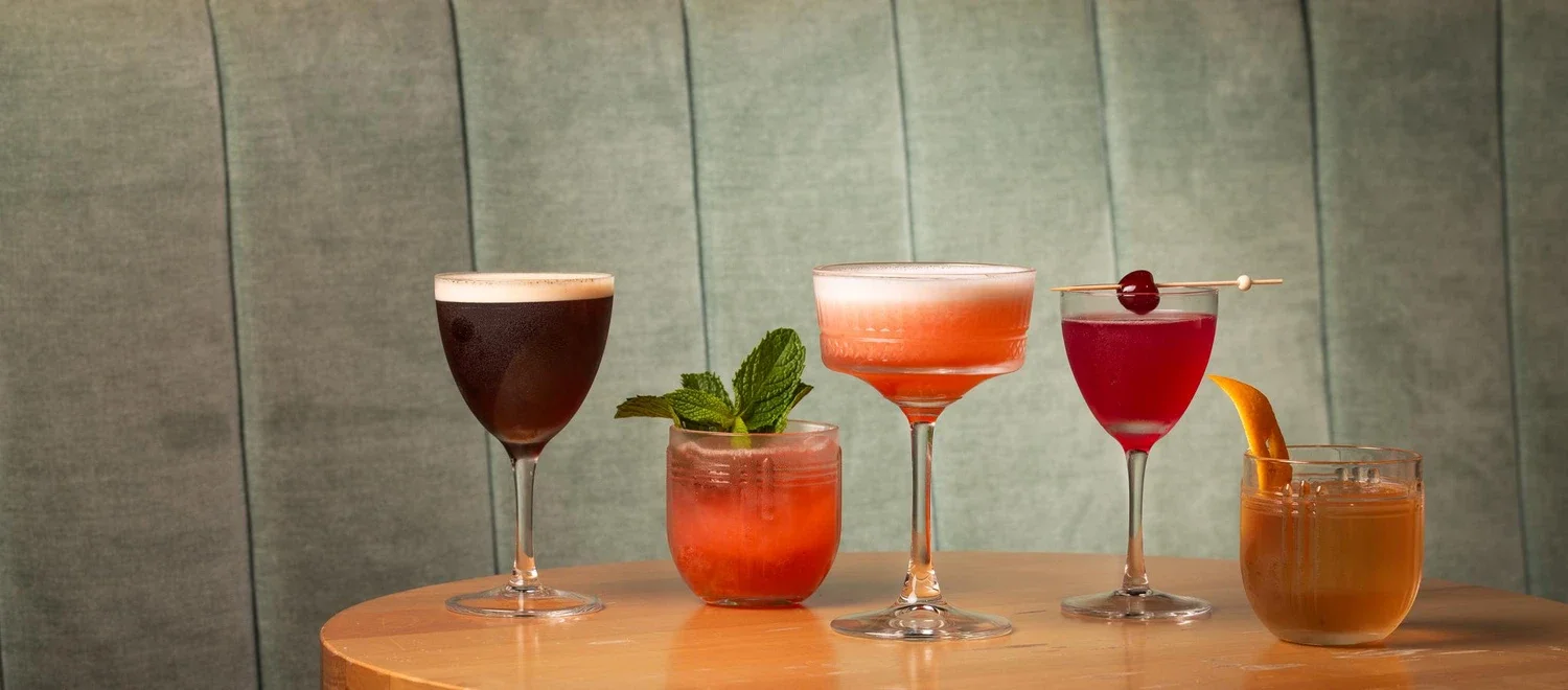 Five cocktails on a wooden table, including a dark beer, a strawberry drink with mint, a pink cocktail in a coupe glass, a red drink with a cherry on top, and an orange drink with an orange slice