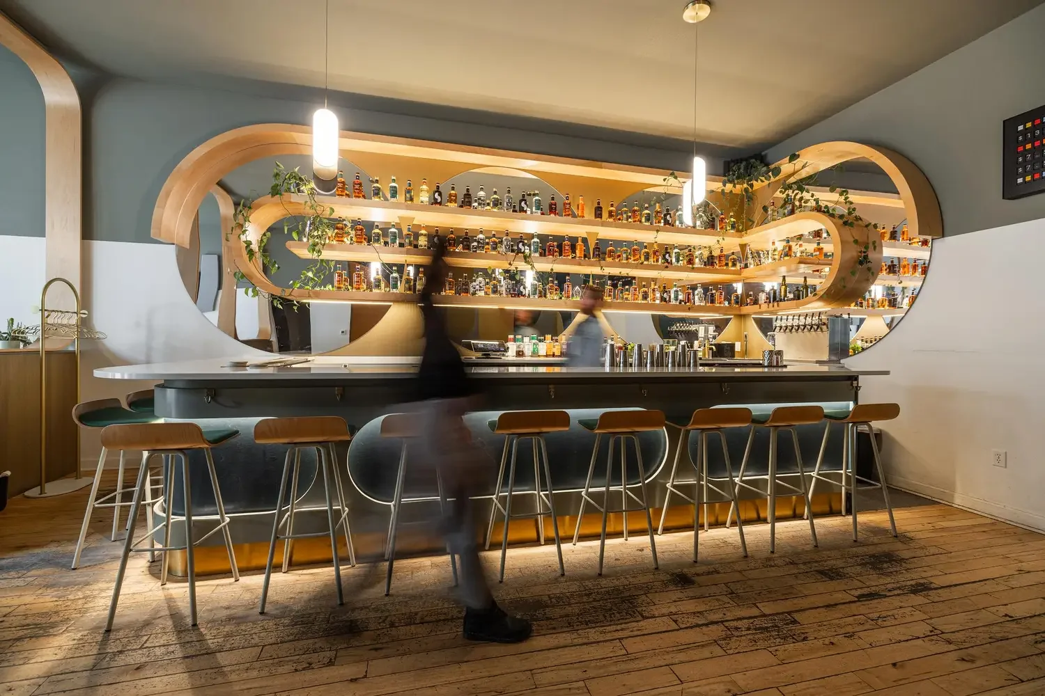 Modern bar with curved wood shelving filled with liquor bottles, a black counter, and bar stools with wood seats, in a well-lit interior.