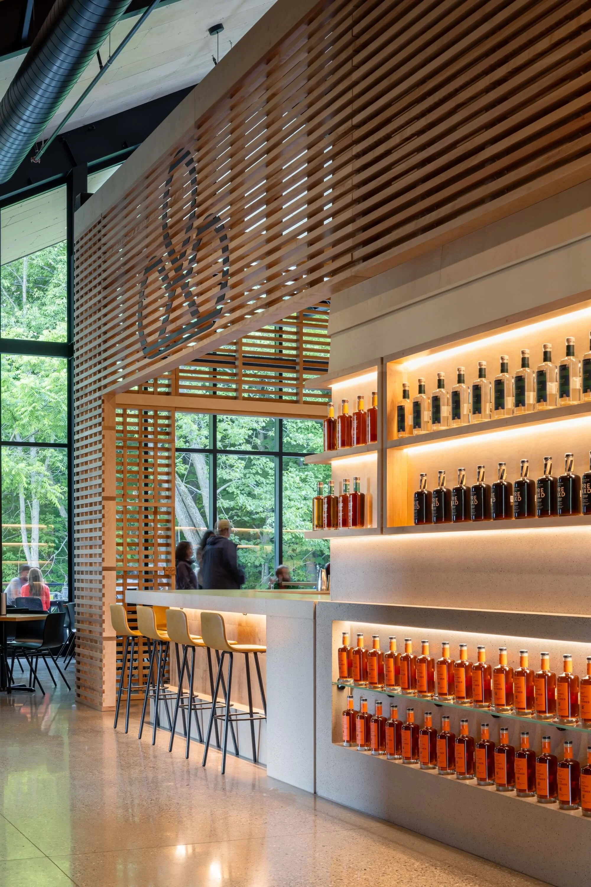 Interior of a modern building with floor-to-ceiling windows, a wooden slatted wall with a circular design, a bar with beige seats, and shelves with orange and black bottles