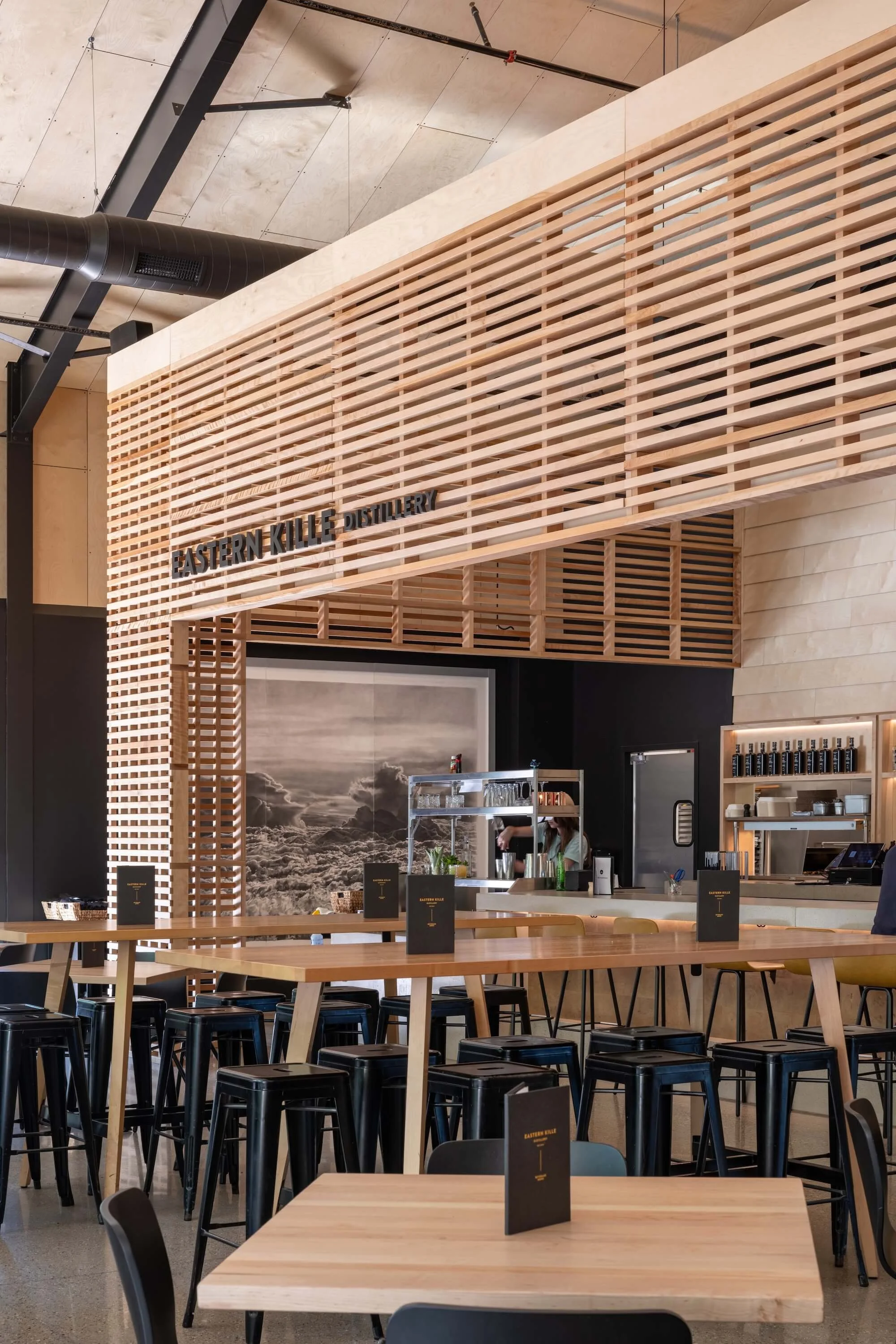 Interior of a modern restaurant or café with wooden tables and black chairs, with a wooden lattice structure overhead displaying the words 'EASTERN KILLE DISTILLERY.' In the background, a barista is preparing drinks behind the counter, which has a large black-and-white mountain scene mural on the wall behind it.