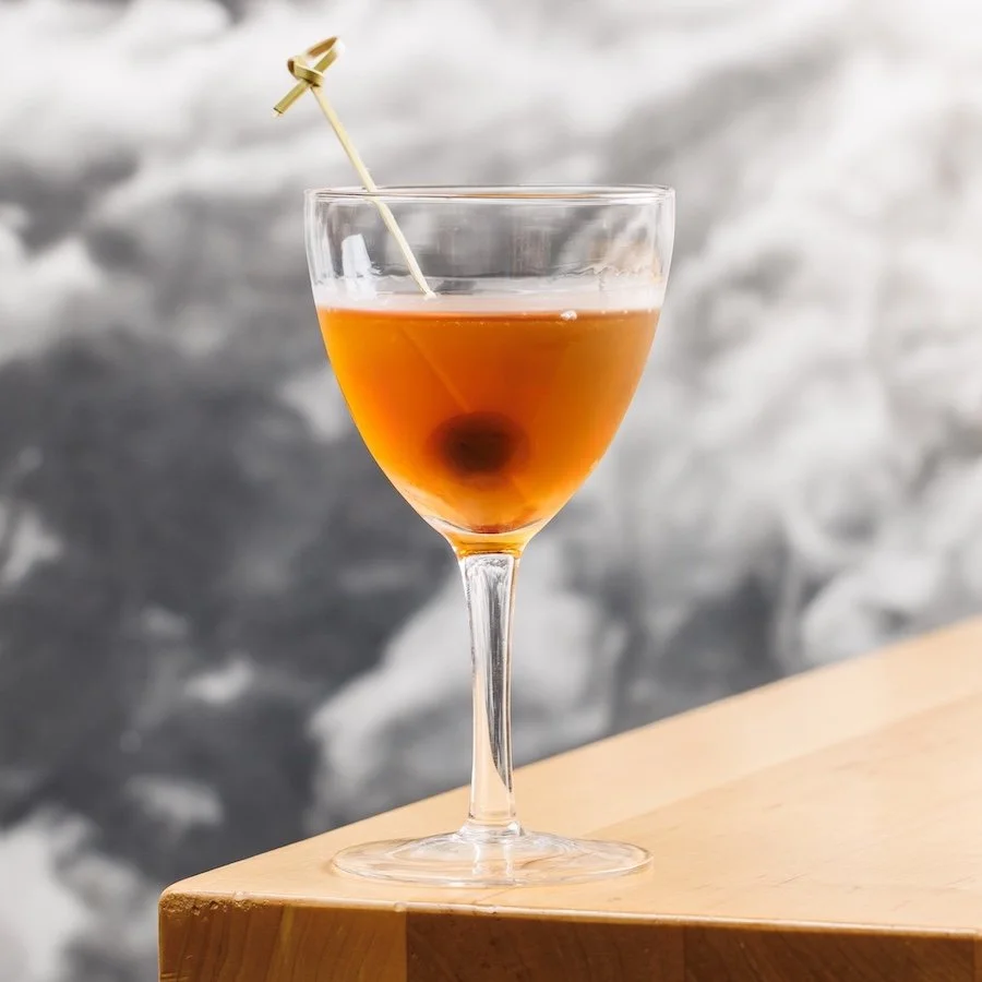 A cocktail in a martini glass with an orange-brown colored liquid and a dark cherry at the bottom, garnished with a decorative cocktail pick.