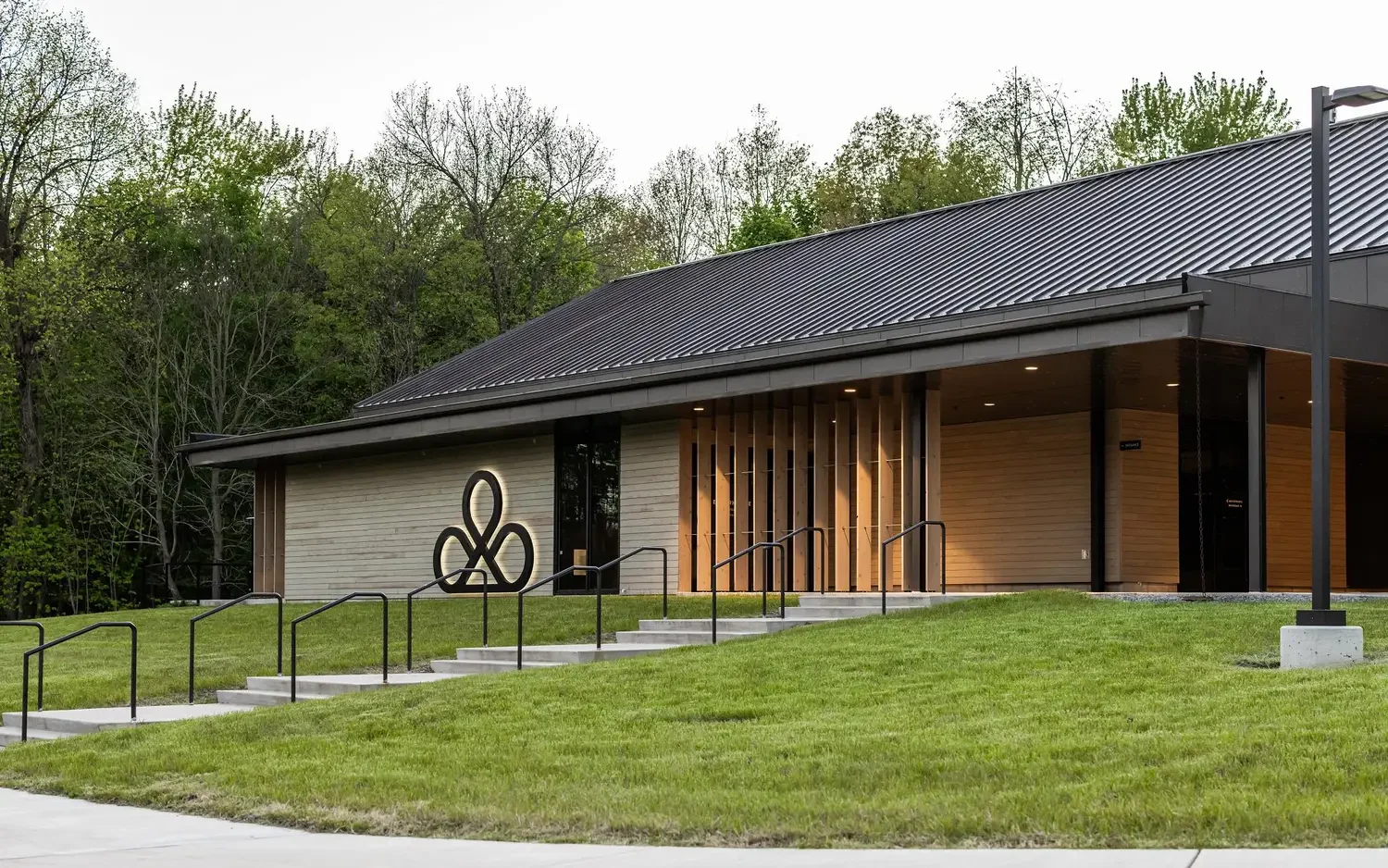 Modern building with wooden exterior, black metal roof, steps, and black metal railing, surrounded by green grass and trees.