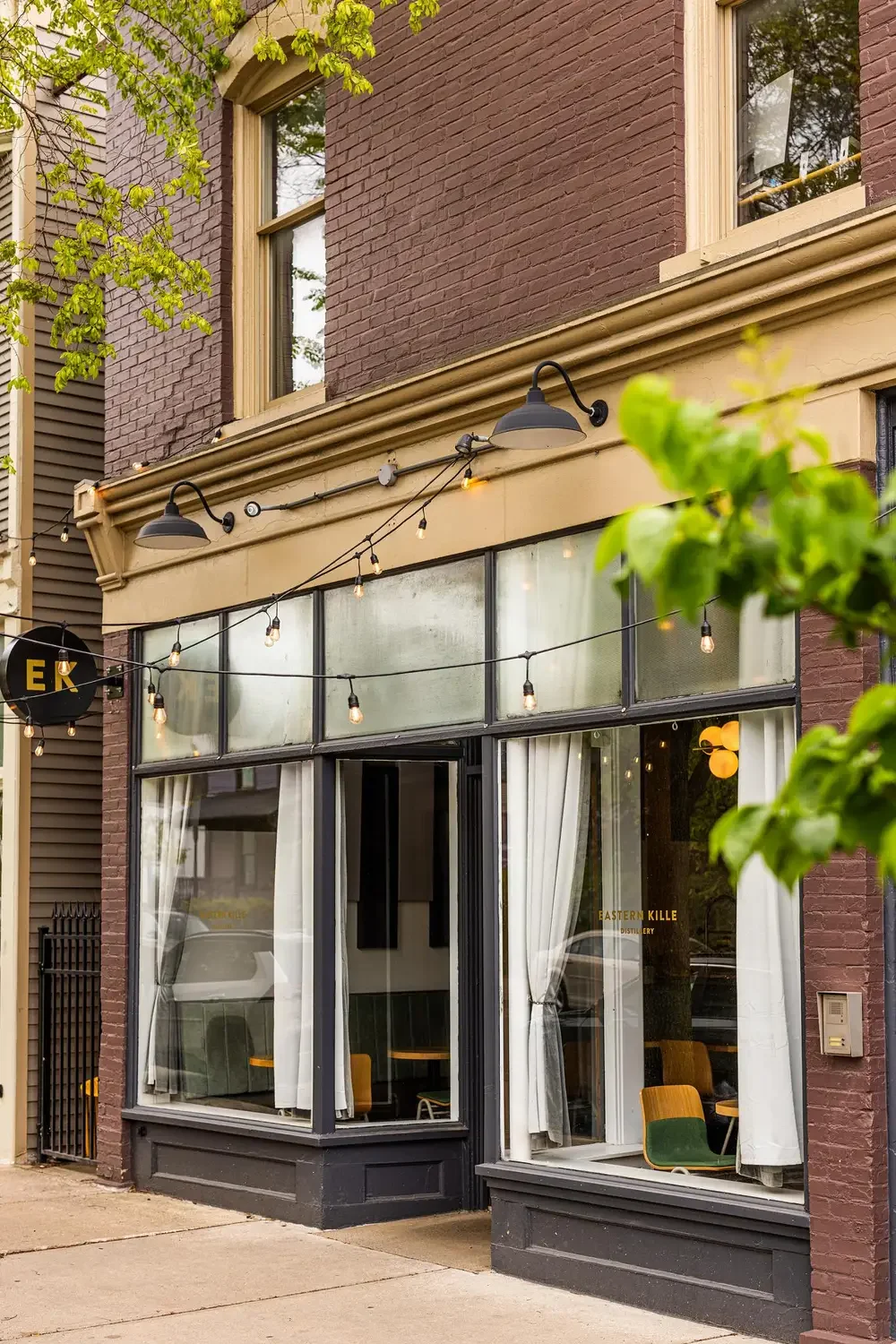 Exterior view of a cafe with large windows, white curtains, and outdoor string lights, brick and beige building facade, and partially visible green trees.