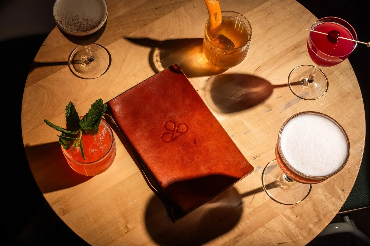 A round wooden table with five colorful cocktails, a brown leather menu with a logo, and soft lighting creating shadows on the table.