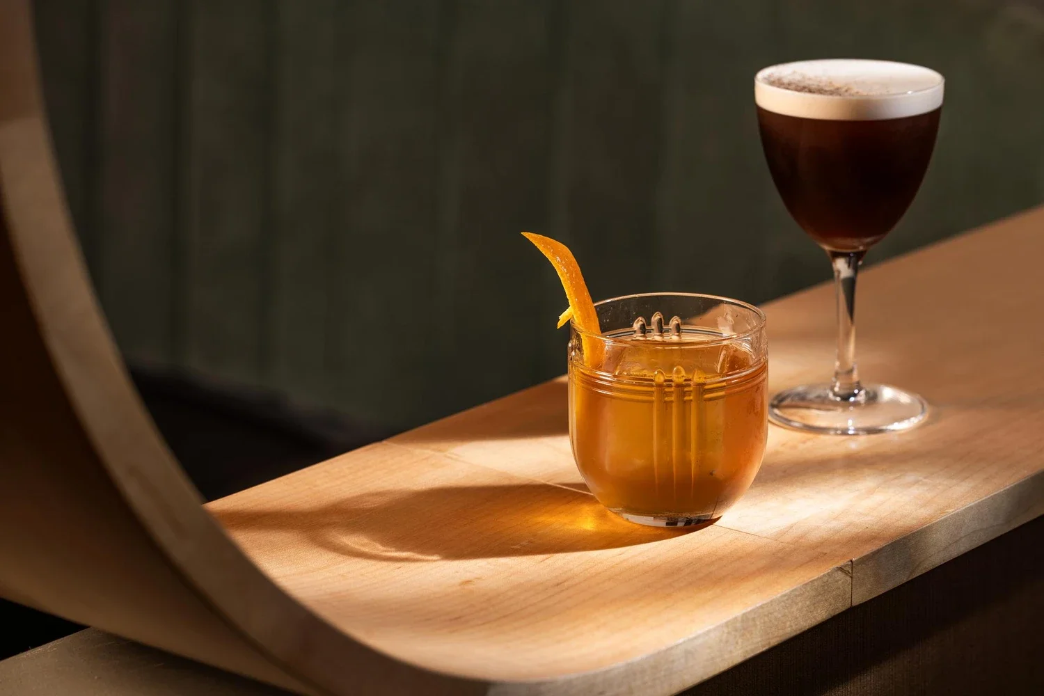 Two cocktails on a wooden tray: a dark cocktail in a champagne flute and an amber-colored drink with an orange twist garnish in a short glass.