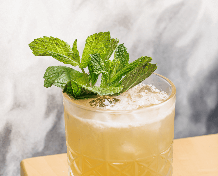 A glass of a yellow-colored beverage garnished with fresh mint leaves on top.