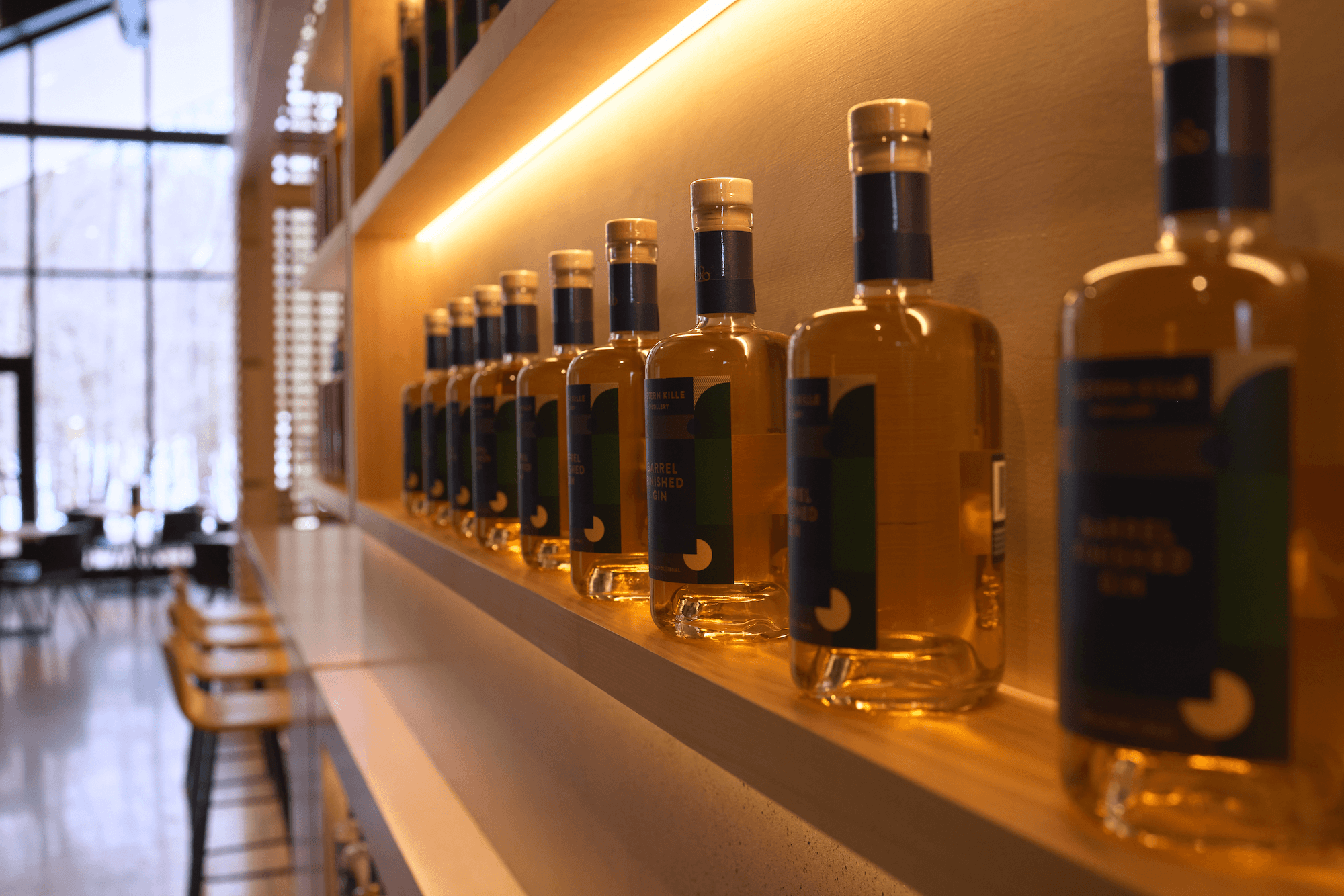 A row of bottles of alcohol displayed on a wooden shelf in a modern, well-lit bar or restaurant with large windows in the background.