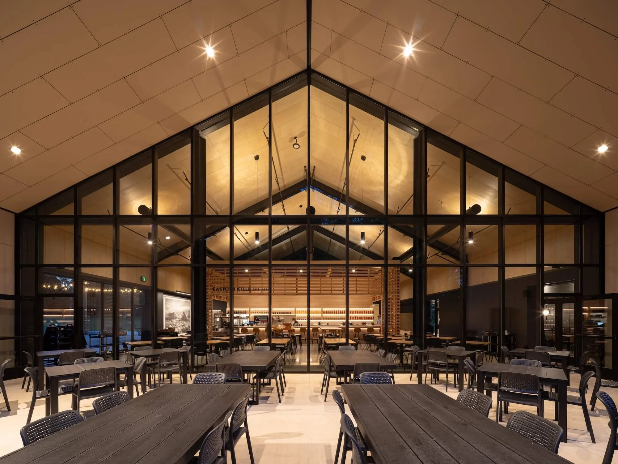 Inside a modern restaurant with a high, peaked ceiling, large glass windows, tables with chairs, and warm lighting.