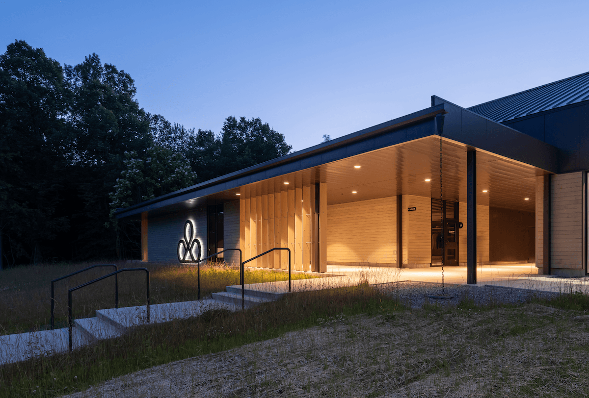 Modern building with a lit entrance, wooden accents, and a logo next to the dark blue exterior, surrounded by trees at dusk.