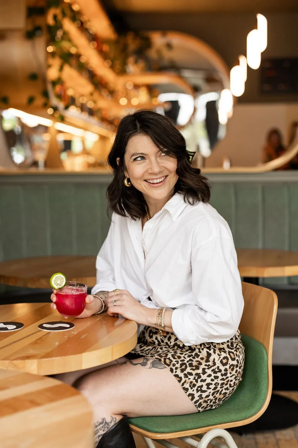 Smiling woman with dark hair, wearing a white blouse and leopard print skirt, sitting at a wooden table in a restaurant, holding a bright pink cocktail with a slice of lime on the rim.