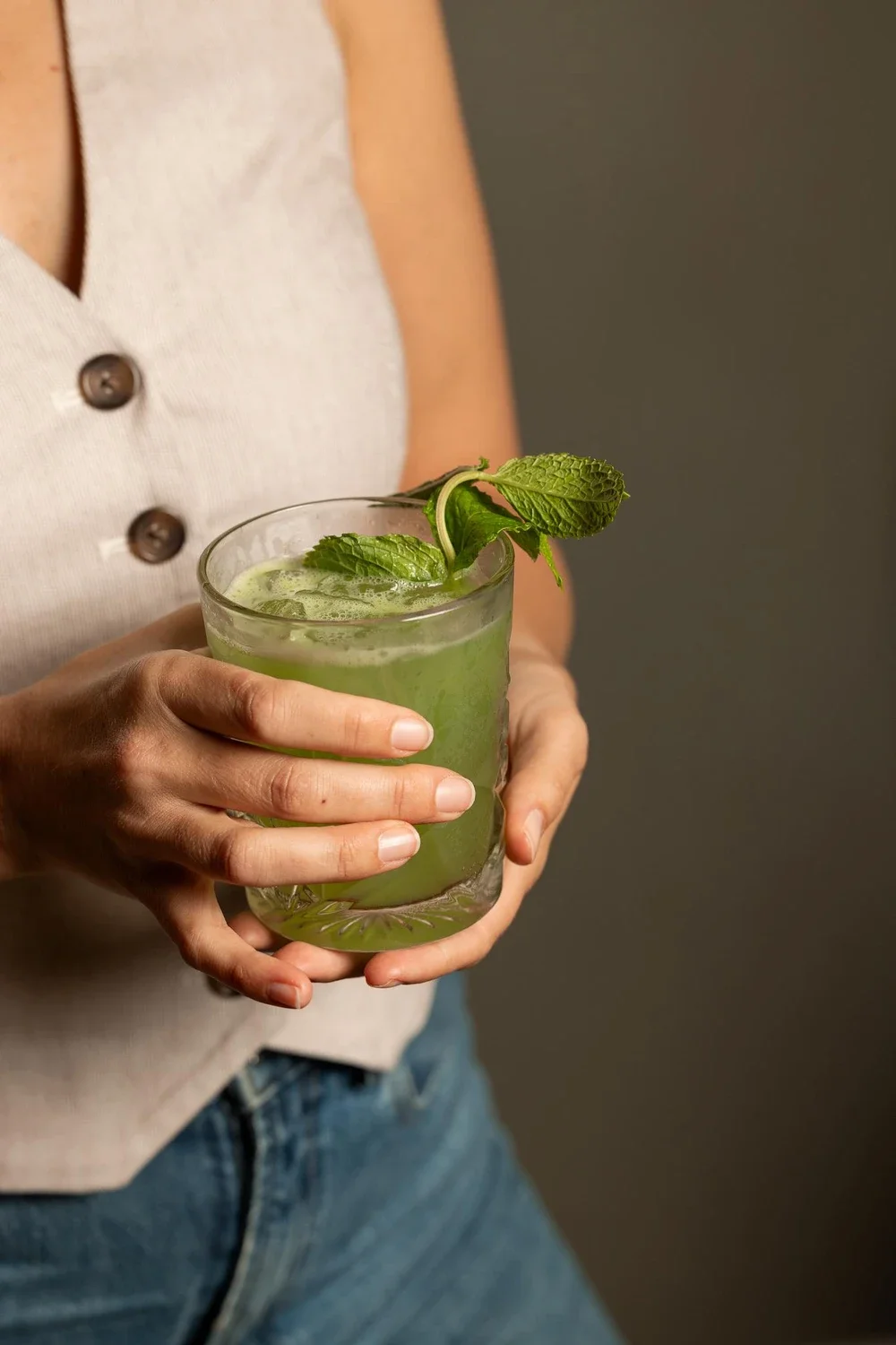 Person holding a tall glass of green cocktail garnished with mint leaves.