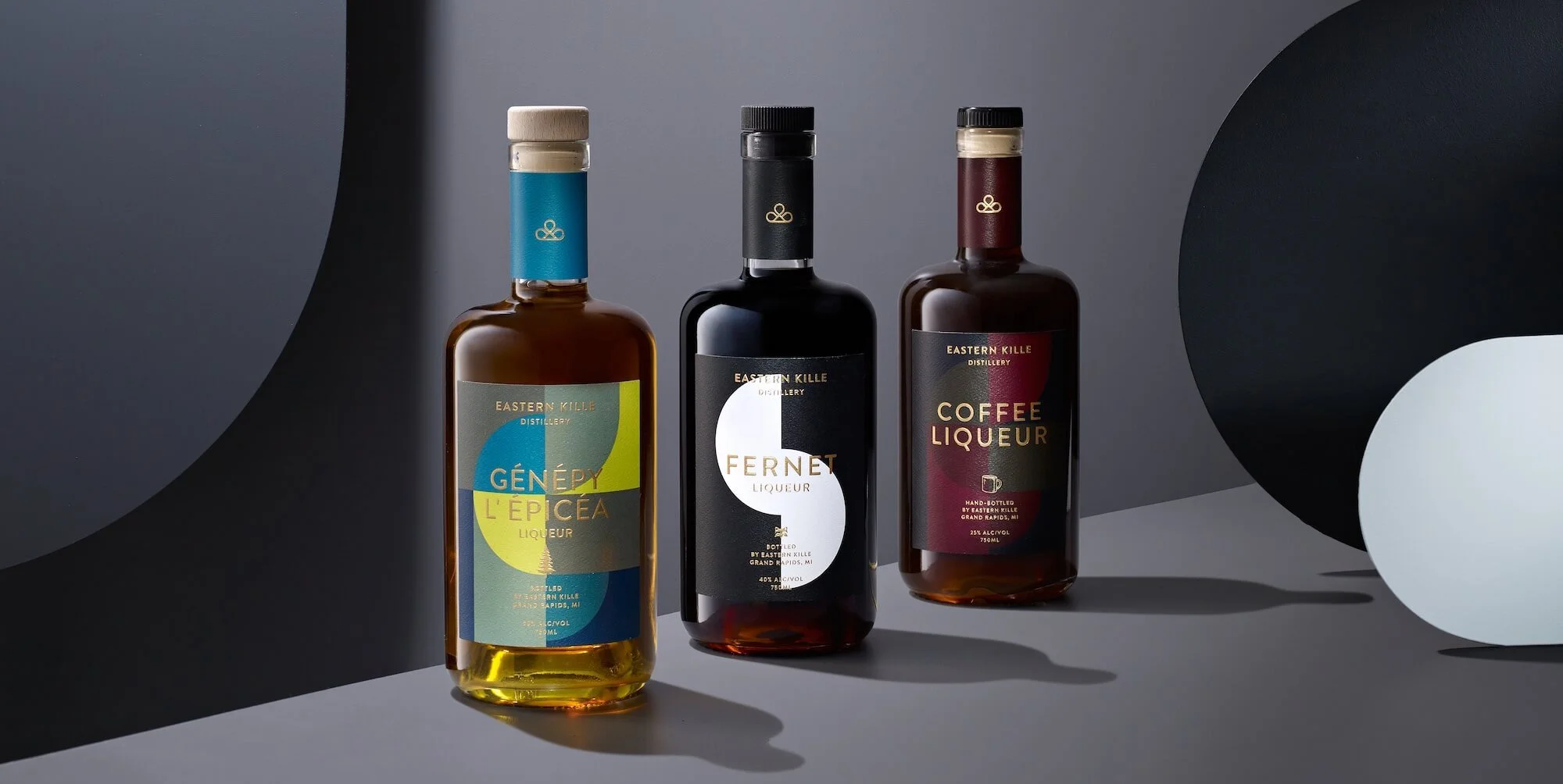 Three bottles of flavored liqueurs from Eastern Kille Distillery are displayed on a gray surface with abstract black and white shapes in the background. The bottles have modern label designs with distinct colors and text.