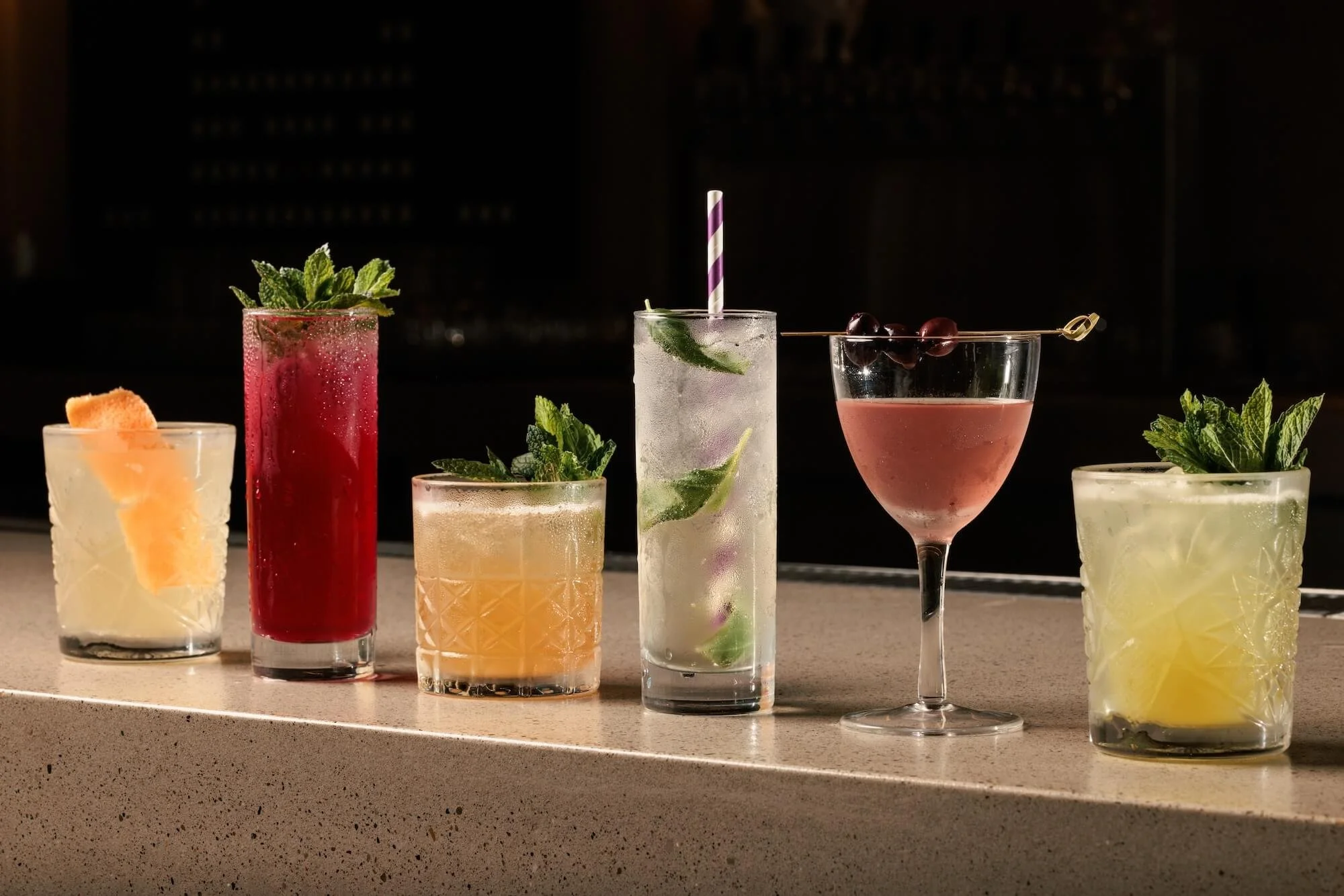 A variety of colorful cocktails on a bar counter, garnished with mint, fruit, and umbrellas.