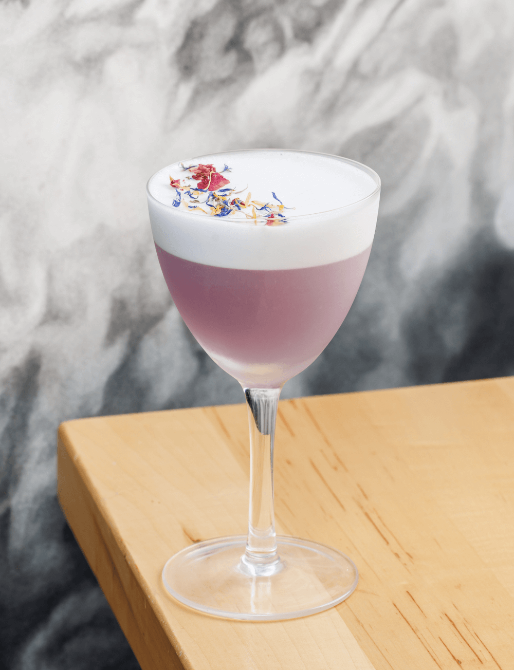 A cocktail in a coupe glass with a pink base, topped with white foam and garnished with colorful edible flowers, placed on a wooden surface against a marbled gray background.