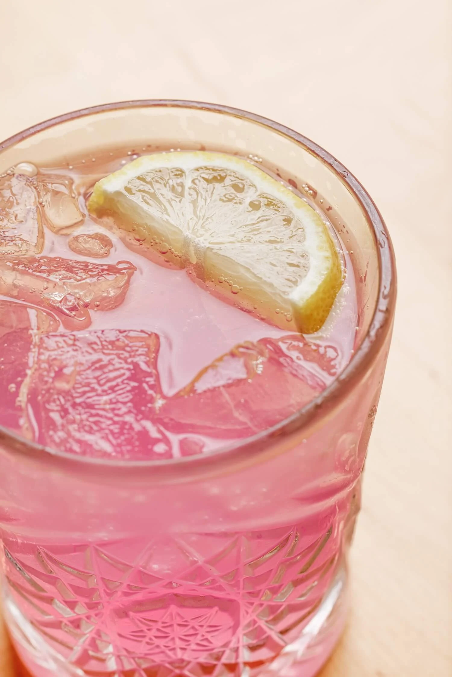 Close-up of a pink glass with a lemon slice and ice cubes inside, filled with pink-colored beverage, on a light background.