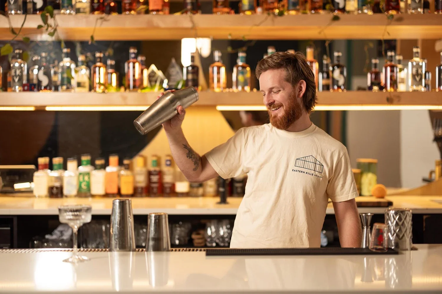 A smiling bartender with a beard and tattooed arm pouring a drink from a metal shaker in a bar with shelves of liquor bottles in the background.