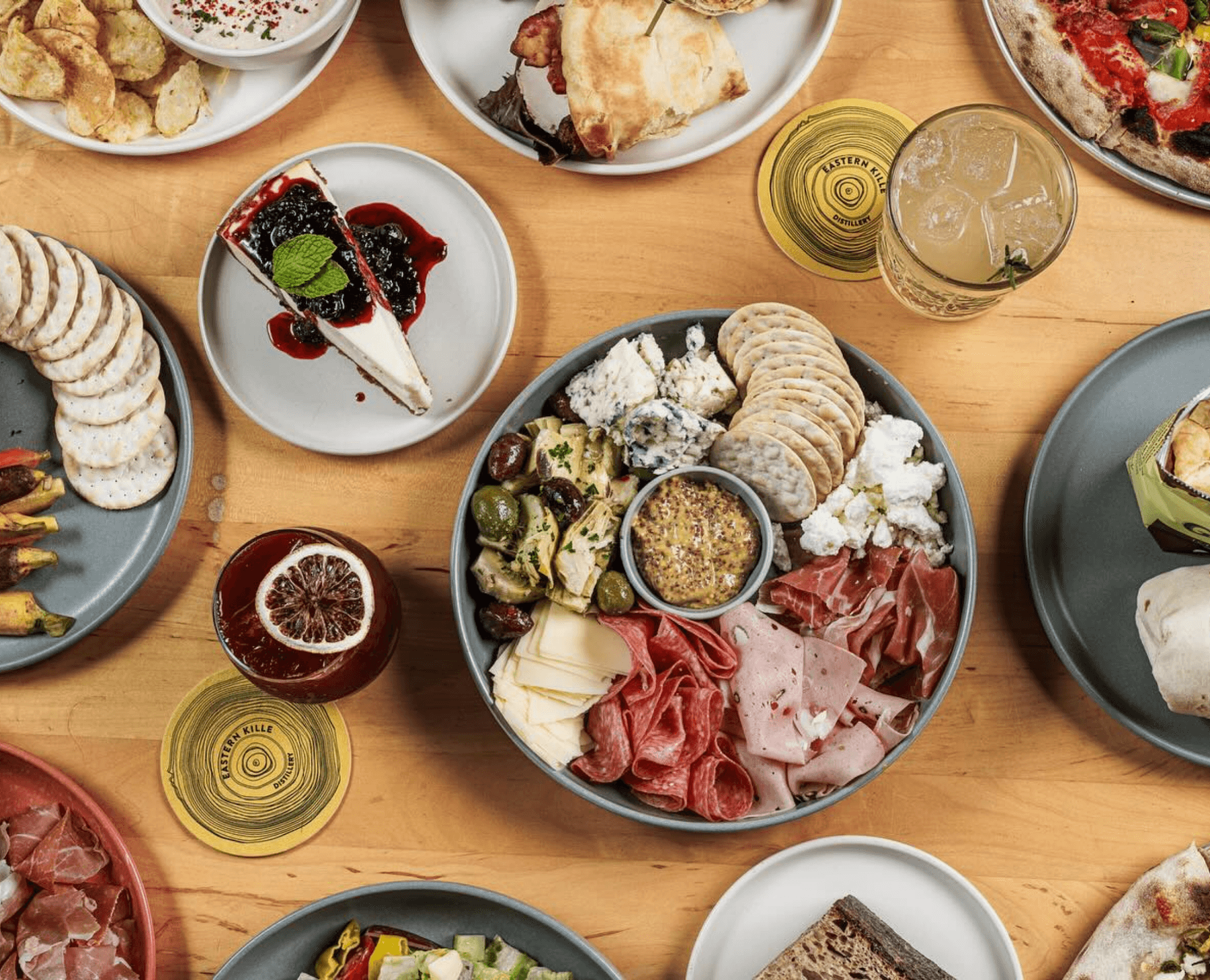 A wooden table holding a variety of appetizers, cheeses, meats, a slice of cheesecake with berry topping, a glass of cocktail, and a slice of pizza.