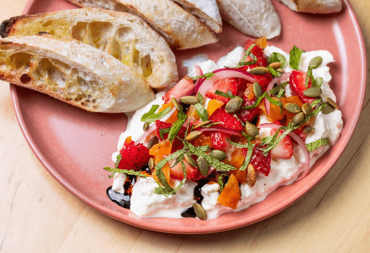 A pink plate with slices of toasted bread and a serving of whipped cottage cheese topped with strawberries, sliced strawberries, chopped herbs, pumpkin seeds, and drizzled with balsamic glaze.