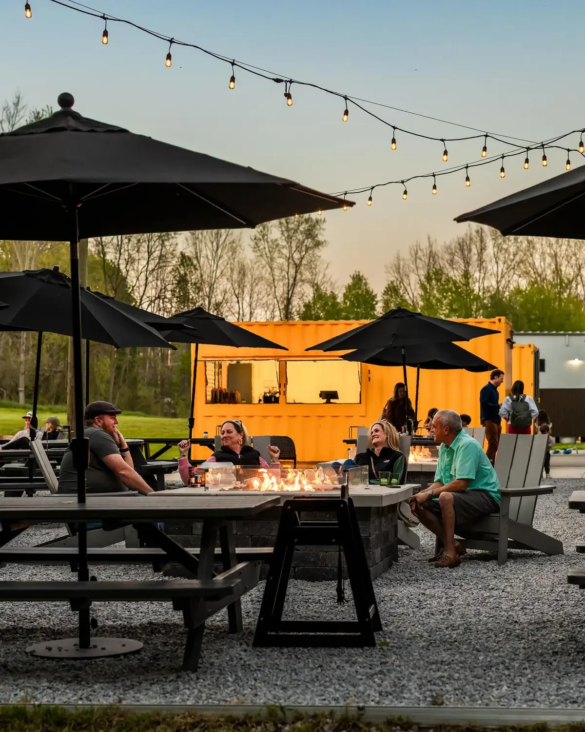 People enjoying outdoor seating at a restaurant with string lights overhead and black umbrellas. There is a yellow building in the background and trees beyond.