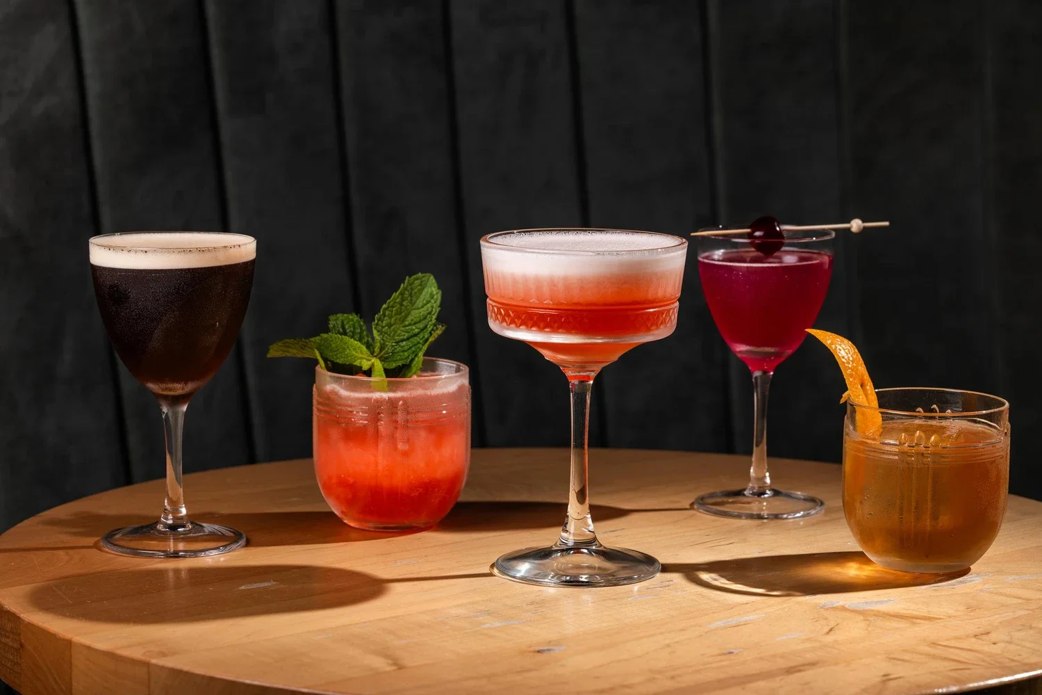 An assortment of five colorful cocktails on a wooden table, with a dark background. The drinks include a black cocktail, a mint garnish on a pink drink, a layered pink and white drink, a bright red cocktail with a cherry garnish, and an orange drink with an orange peel.