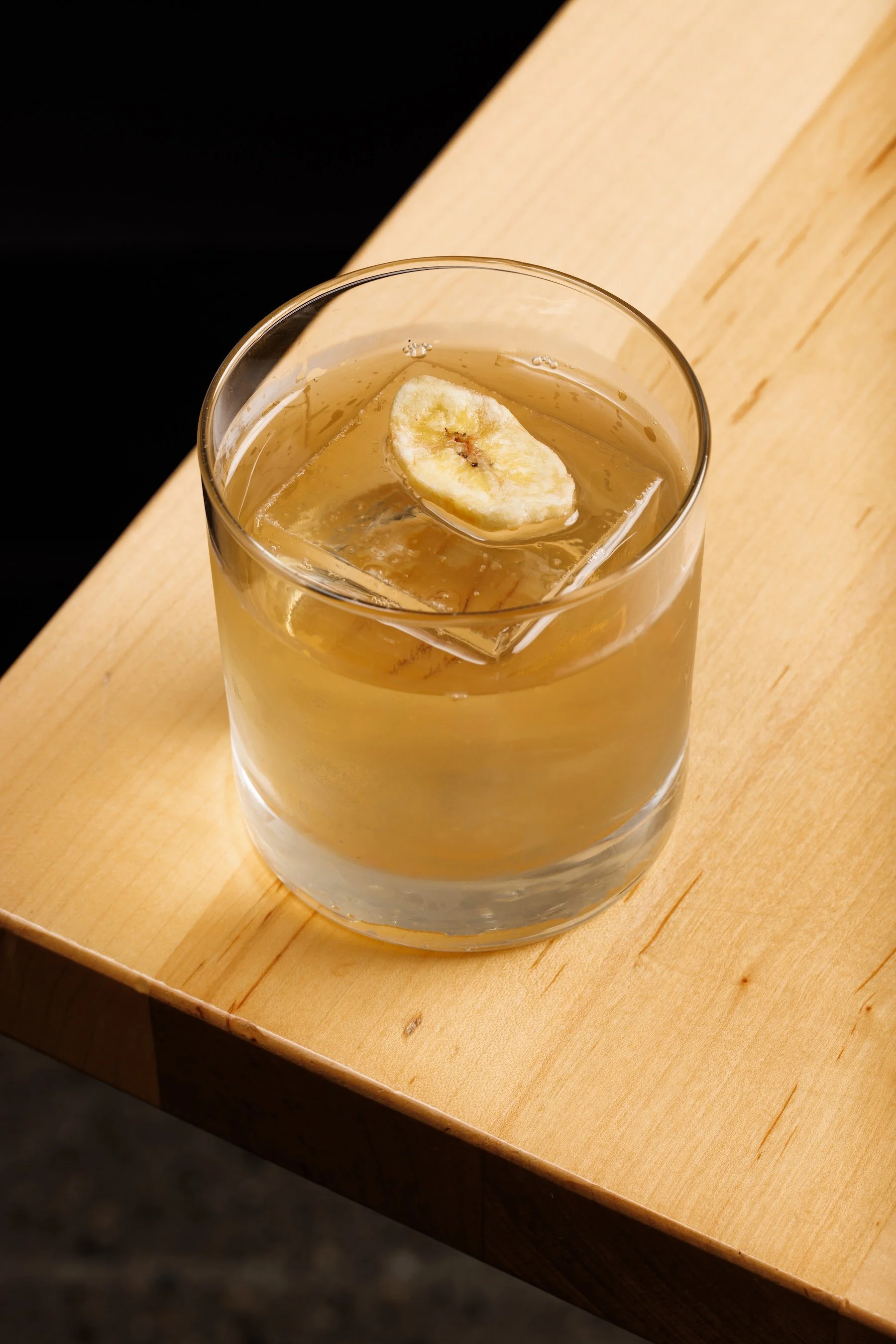 A clear glass filled with a golden-colored drink and ice cubes, garnished with a slice of banana, placed on a wooden surface.