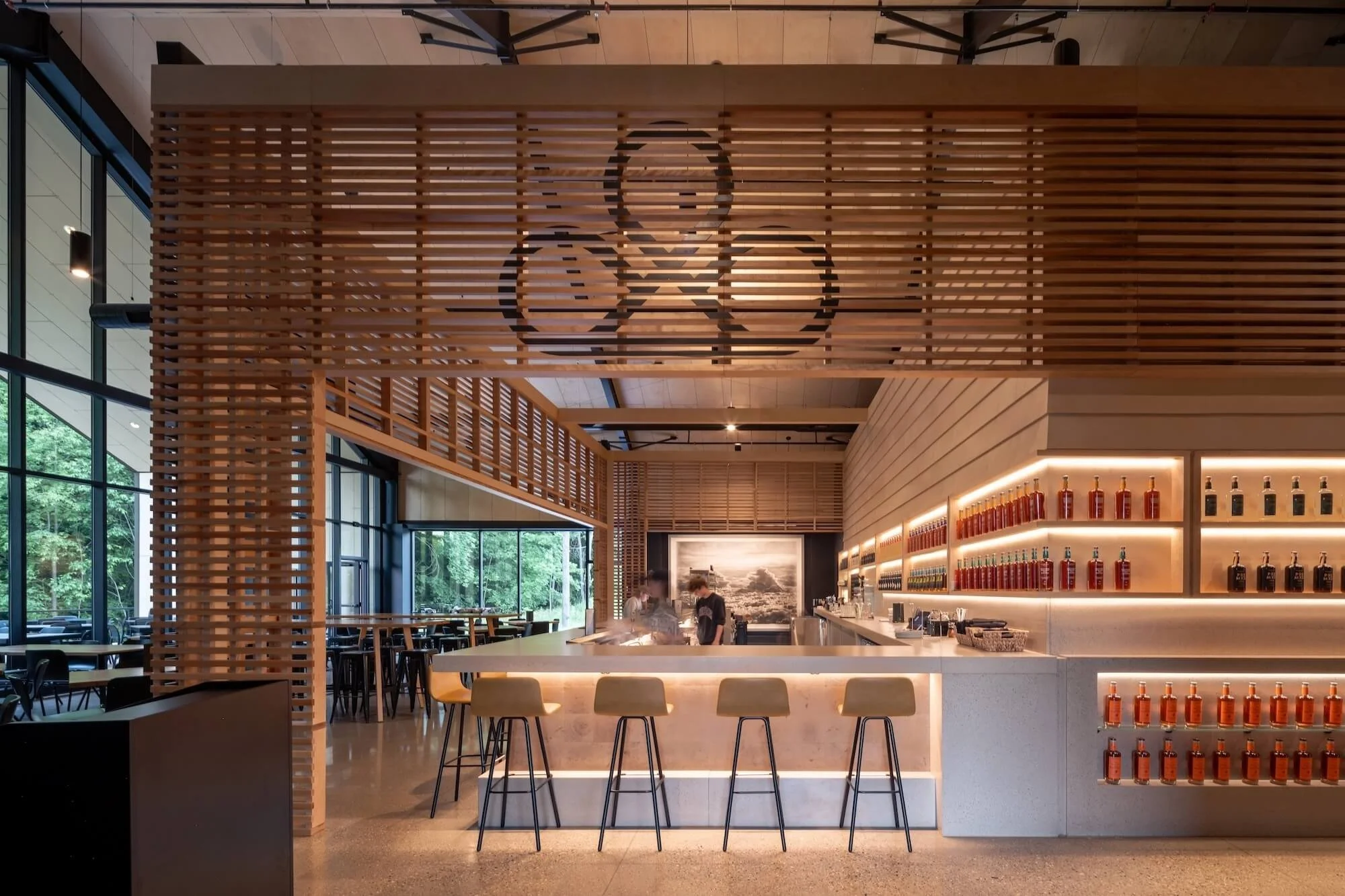 Interior of a modern café or restaurant with wooden accents, a bar area with bottles on shelves, and large windows with outside greenery visible.