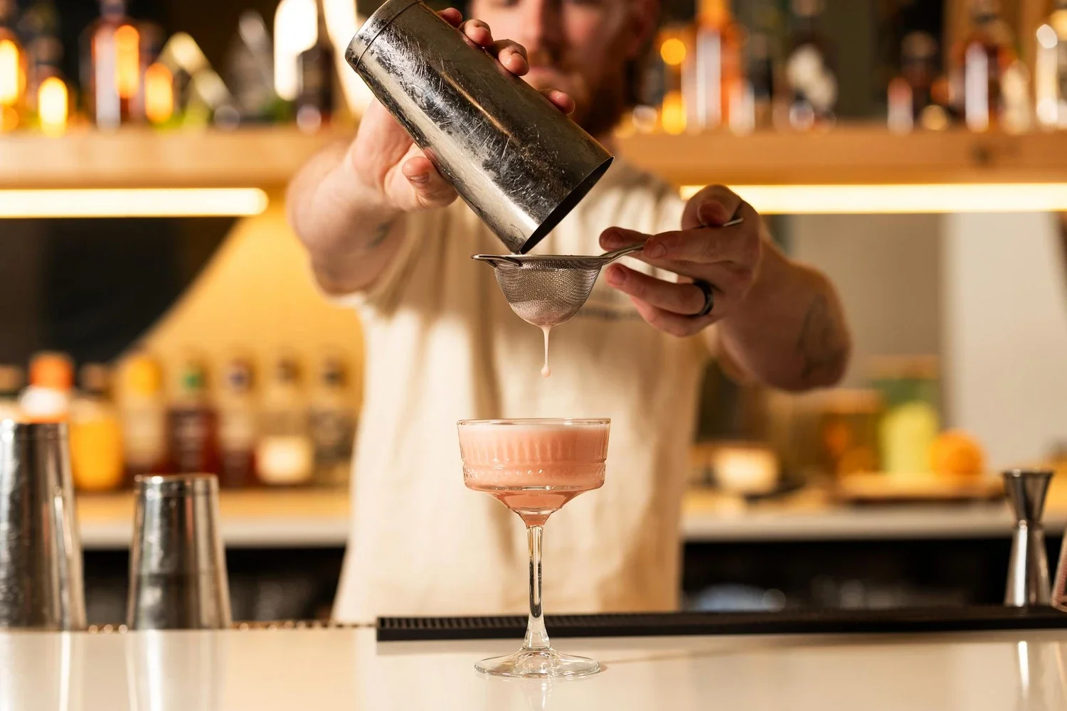 Bartender pouring pink cocktail through a fine strainer into a coupe glass