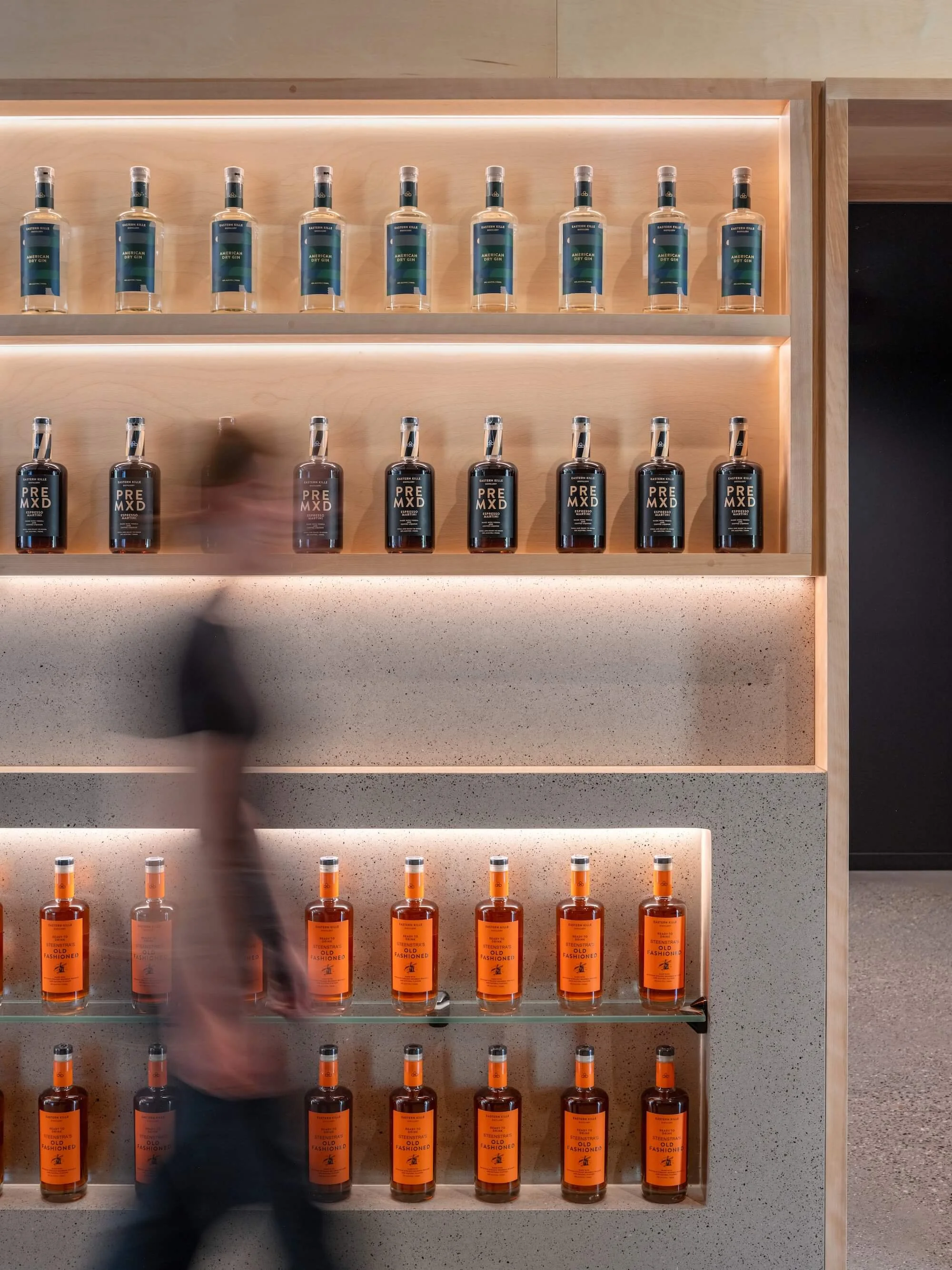 Display of spirits bottles on wooden shelves, with a blurred person walking past in foreground.