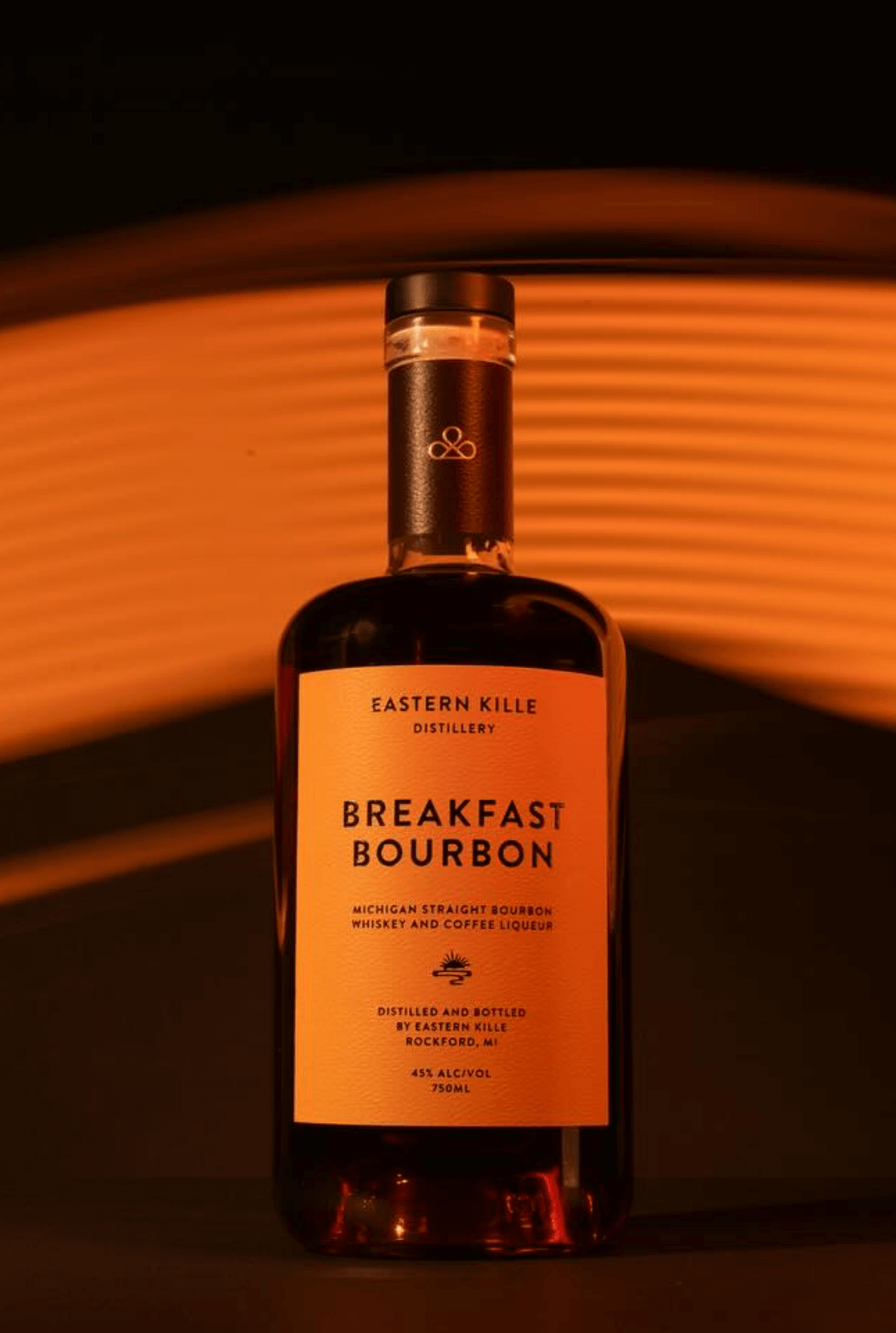 A bottle of Eastern Kille Distillery Breakfast Bourbon Michigan straight bourbon whiskey and coffee liqueur with an orange label, set against a background of horizontal orange light stripes.