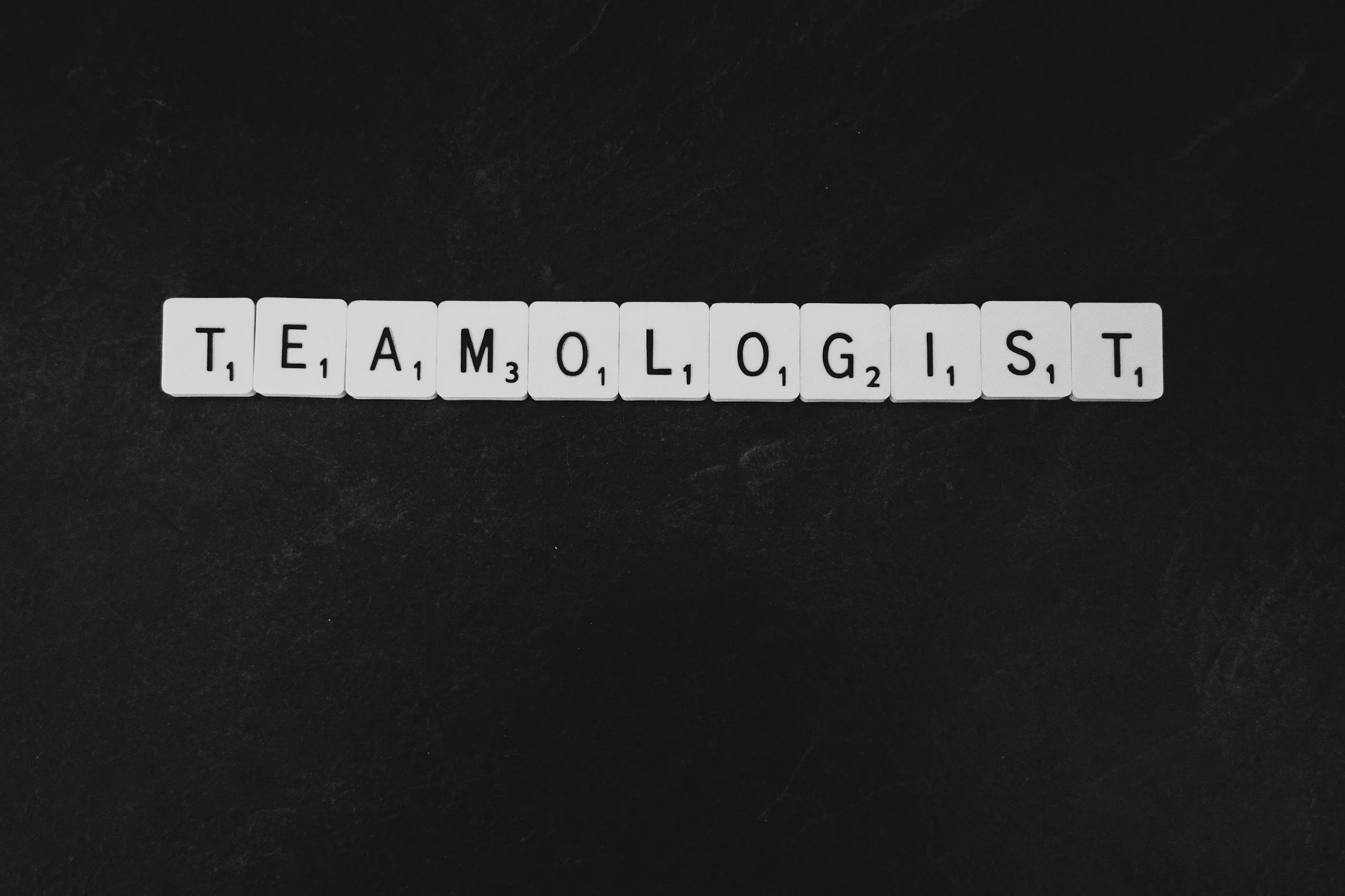 Scrabble tiles spelling 'TEAMOLOGIST' on a black surface.