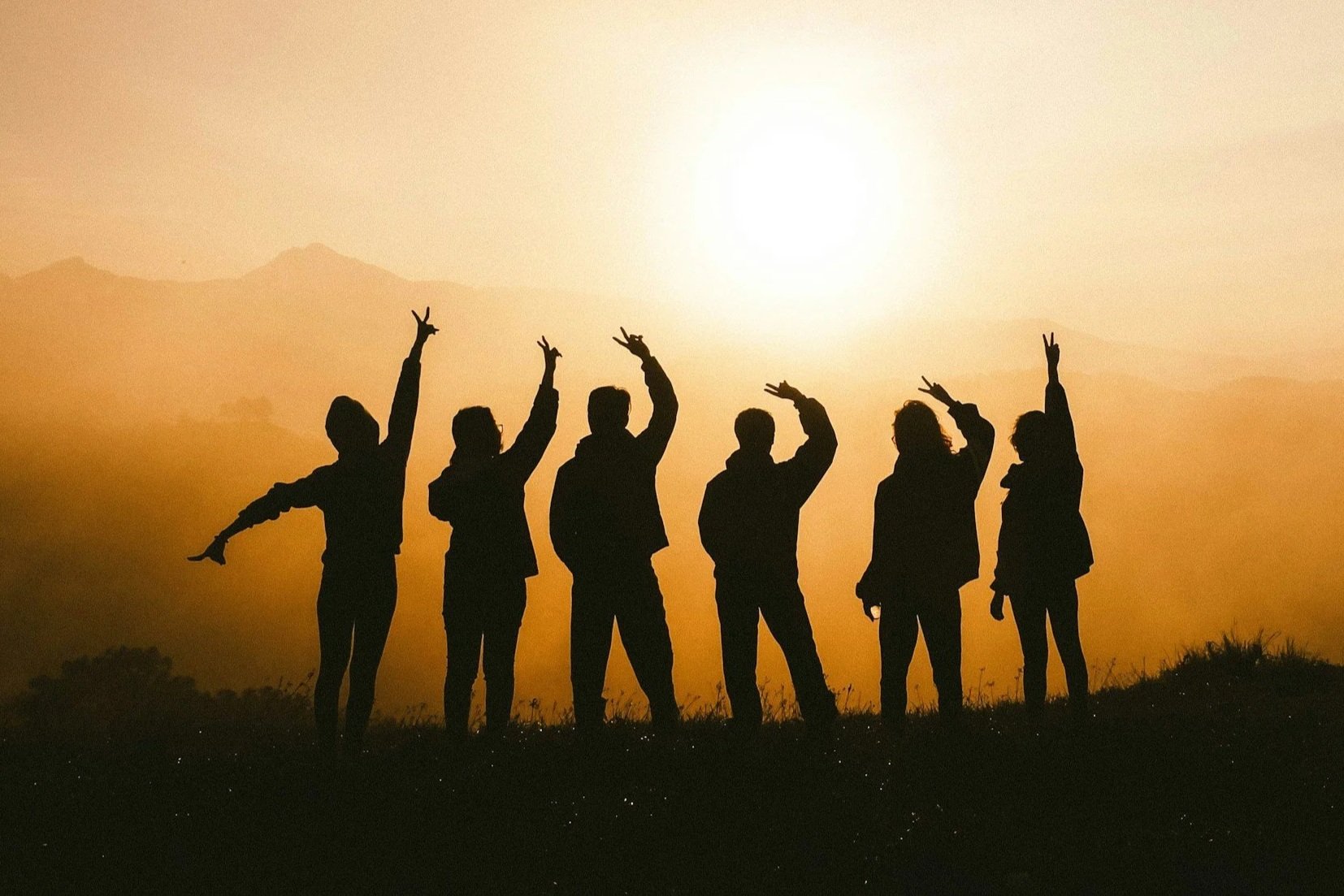 Silhouettes of six people standing on a hill during sunset, making peace signs and playful gestures.