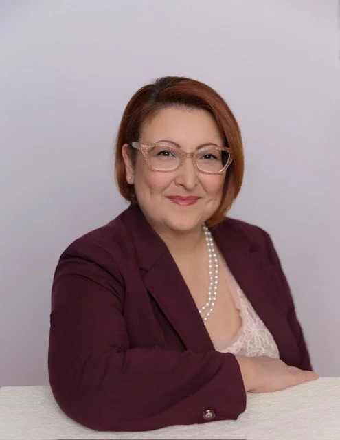 Headshot of a woman with short red hair, glasses, wearing a purple blazer and pearl necklace, smiling at the camera.