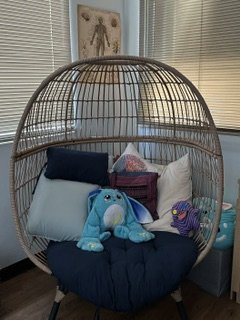 Rattan hanging pod chair with assorted pillows and stuffed toys inside, situated near windows with blinds.