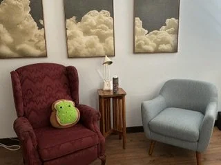 Living room with a red armchair, a light gray armchair, a wooden side table, and three cloud-themed artwork pieces on the wall. There is a green plush toy on the red armchair.