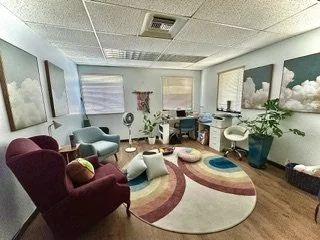 A cozy office lounge with colorful chairs, a patterned rug, potted plants, and large cloud-themed artwork on the walls.