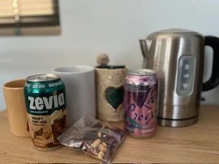A countertop with a can of Zevia coffee, a white mug, an open bag of snacks, a pink beverage can, and a stainless steel electric kettle.